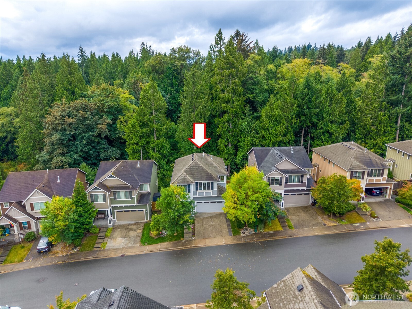 Aerial view of a gray two-story house marked by a red arrow, with evergreen forest behind it.