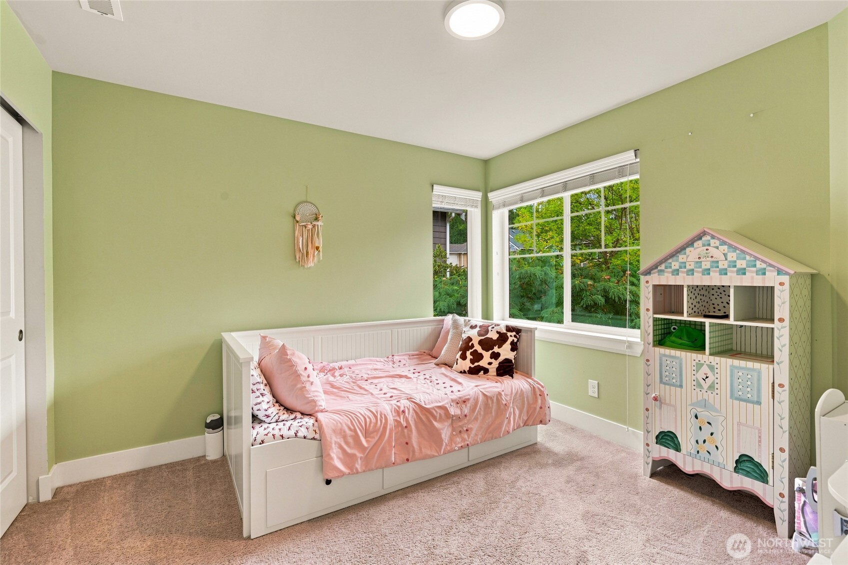 Furnished bedroom with a pink-dressed daybed, green walls, large windows, and a decorative dollhouse.