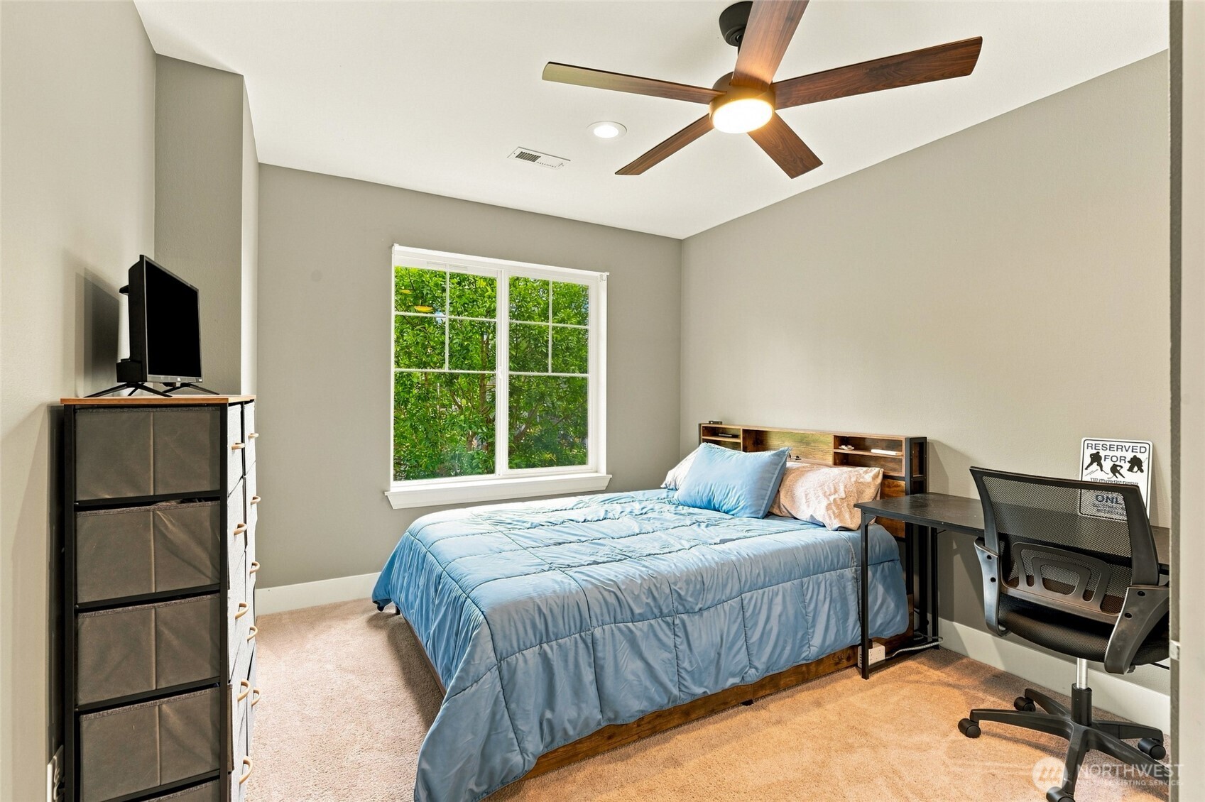 Bedroom with a blue quilted bed, wood headboard, desk, and window overlooking green foliage.