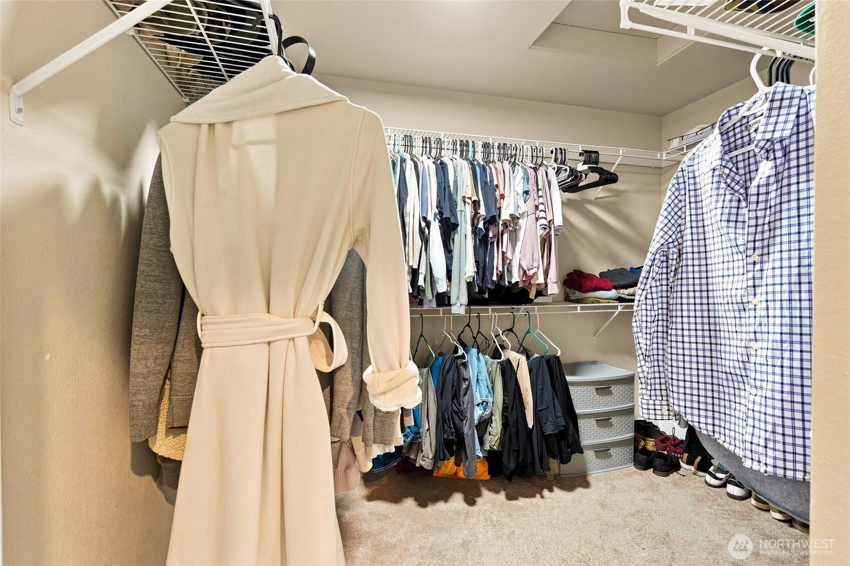 Walk-in closet with white wire shelving, hanging clothes, carpet, and storage drawers.