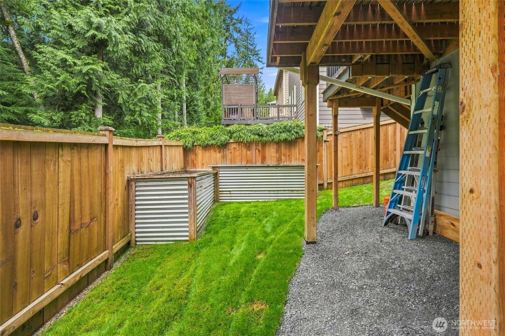 Fenced backyard with a covered wooden structure, green lawn, gravel walkway, and corrugated metal garden beds.
