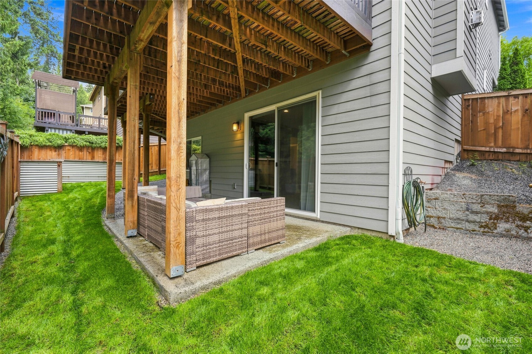 Covered patio beneath a raised wooden deck, with wicker seating beside the home and a fenced lawn.