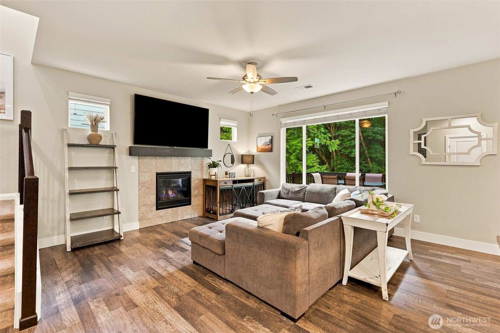 Staged living room with a gray sectional, tiled fireplace, and sliding doors overlooking trees.