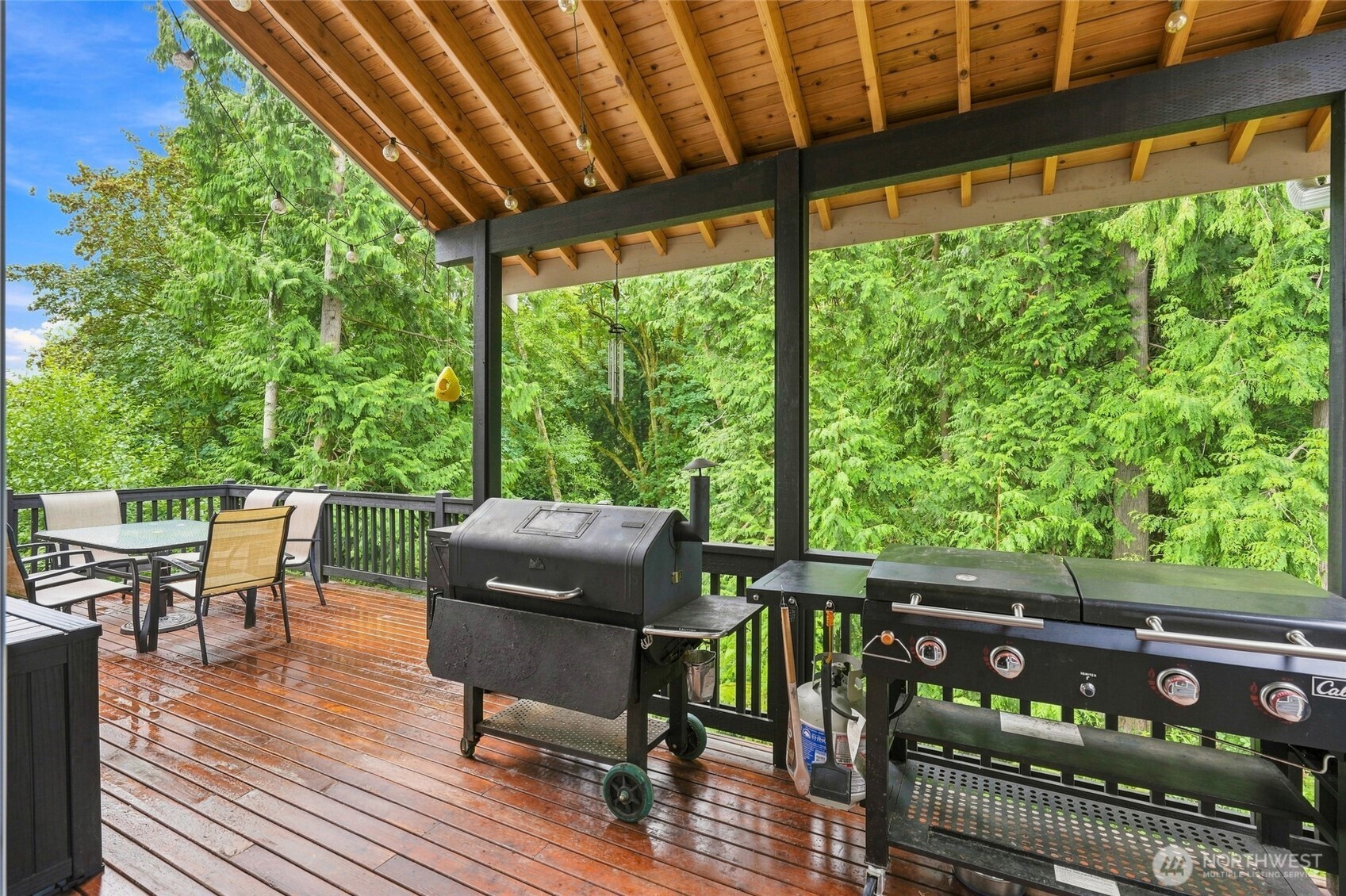 Covered deck with dining furniture and two grills overlooking a lush forest.