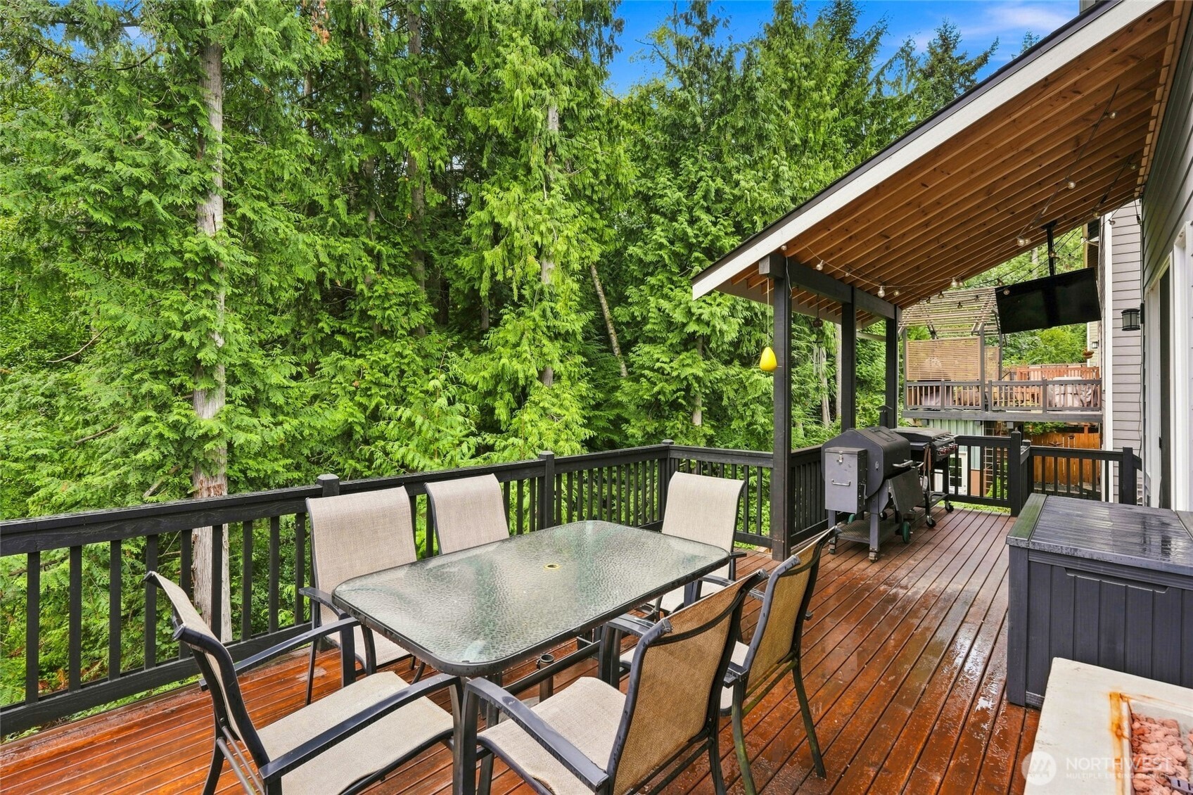 Covered wooden deck with dining set, grill, and hot tub overlooking evergreen trees.