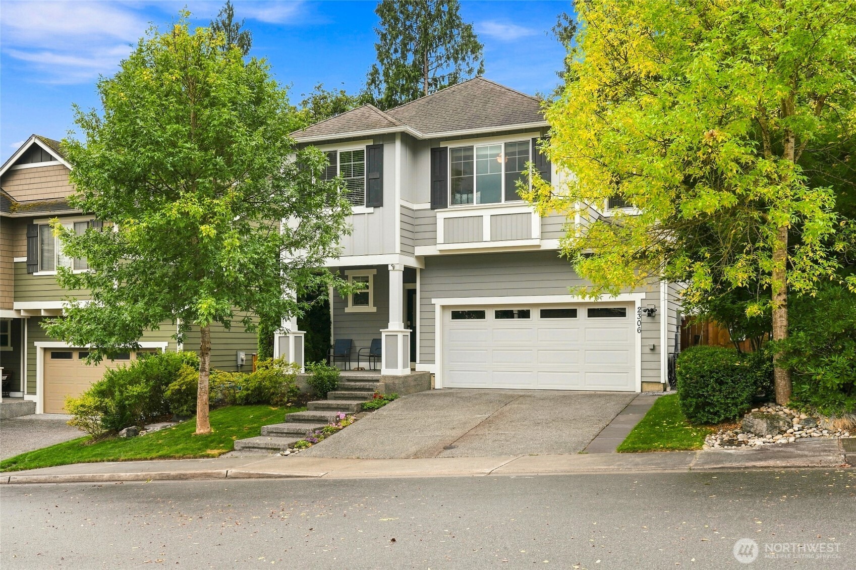 Two-story gray house with a double garage, front steps, and mature trees framing the facade.