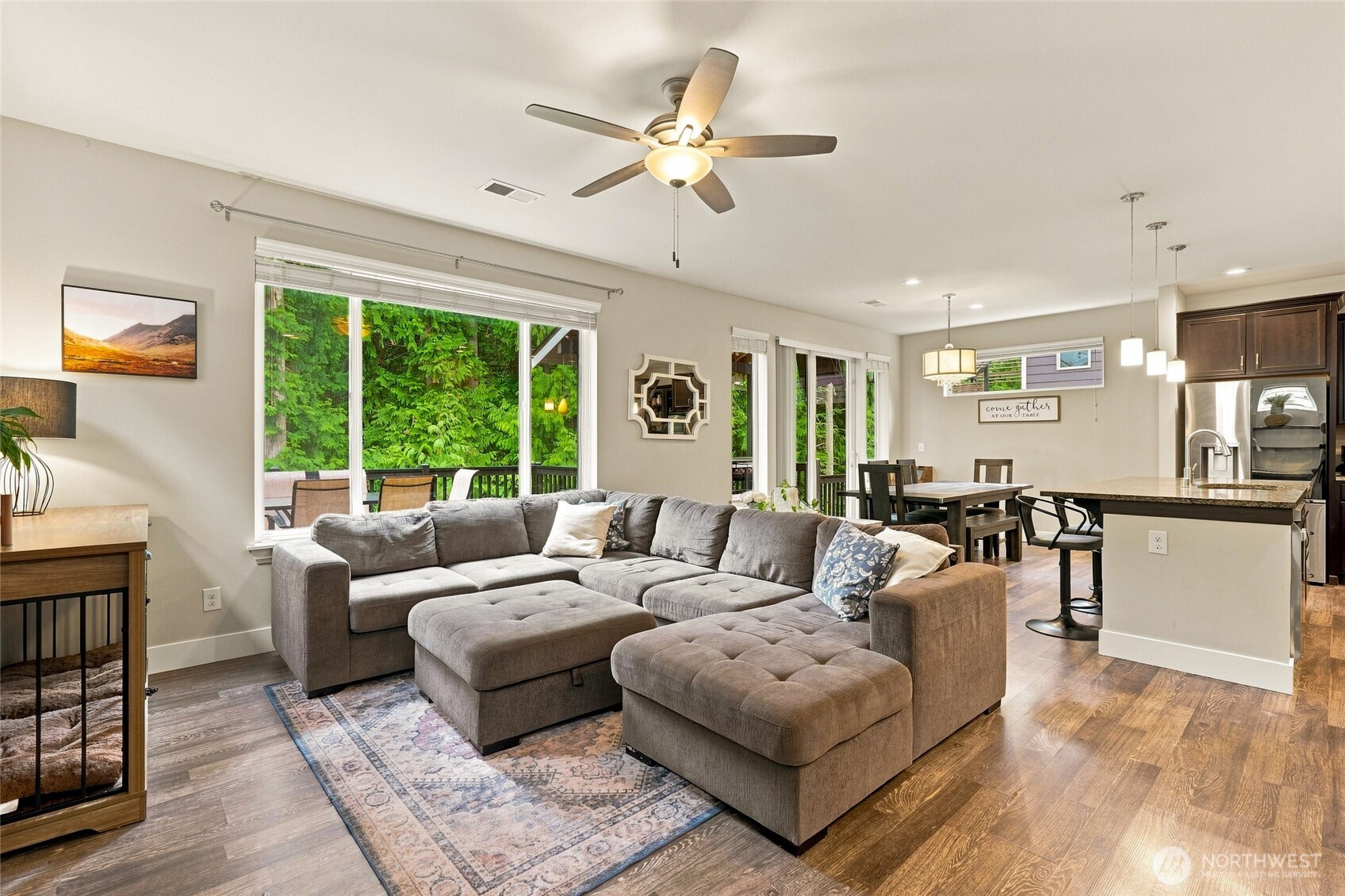 Open-plan living room with a large gray sectional, dining table, kitchen island, and leafy backyard views.