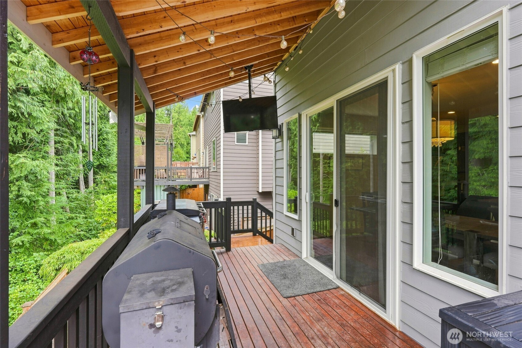 Covered wooden deck with a large barbecue grill, mounted TV, and wooded surroundings.