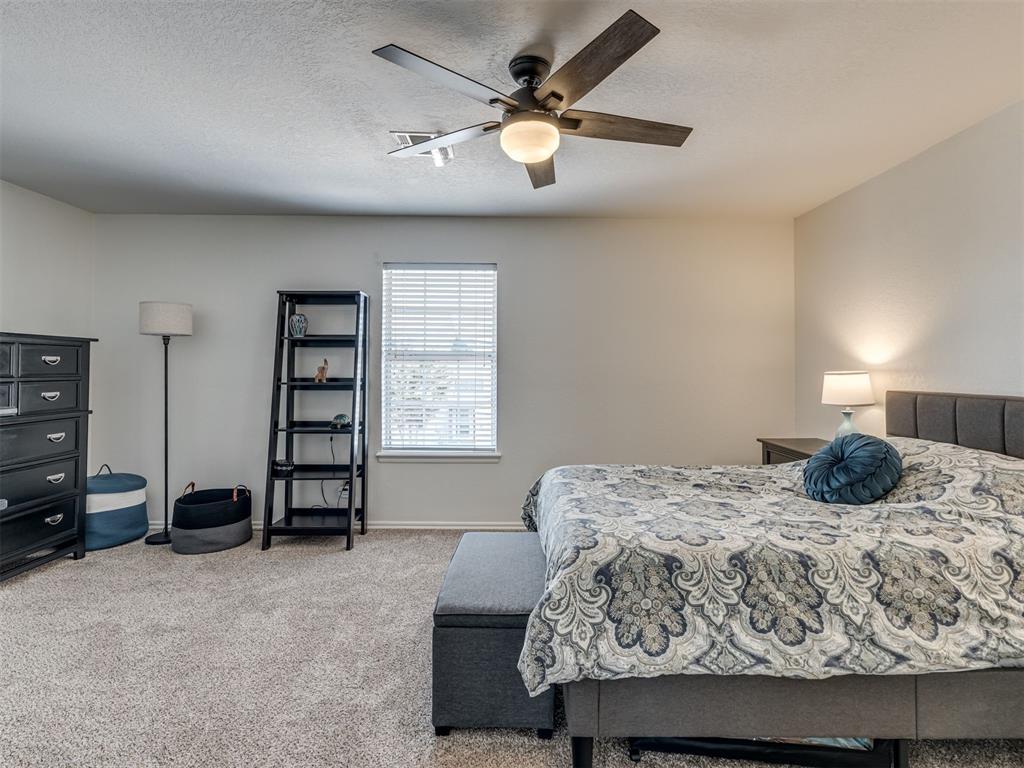 Furnished bedroom with a patterned bedspread, carpet, window blinds, and dark ceiling fan.