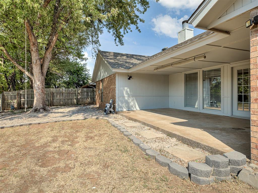 Covered backyard patio beside a brick house, with large windows, a mature shade tree, and a fenced yard.