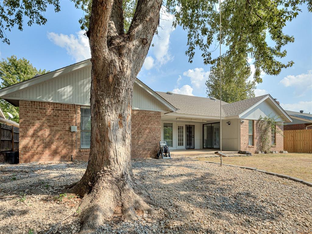 Brick house with a covered patio and gravel backyard, partly obscured by a large mature tree.