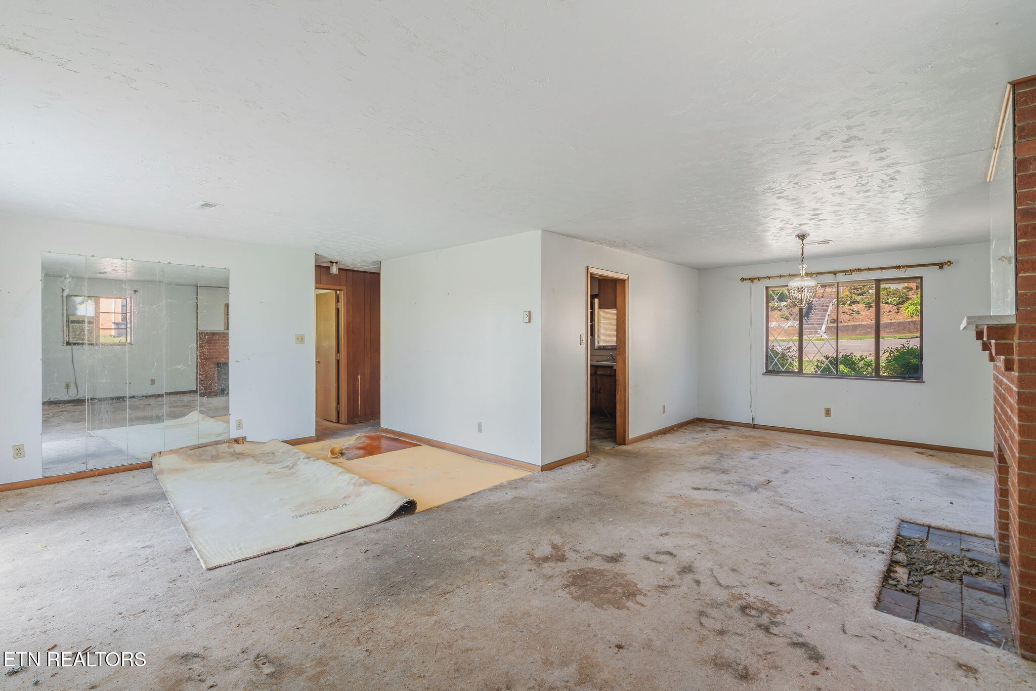 Vacant living and dining room with a brick fireplace, mirrored wall, and sections of carpet removed.