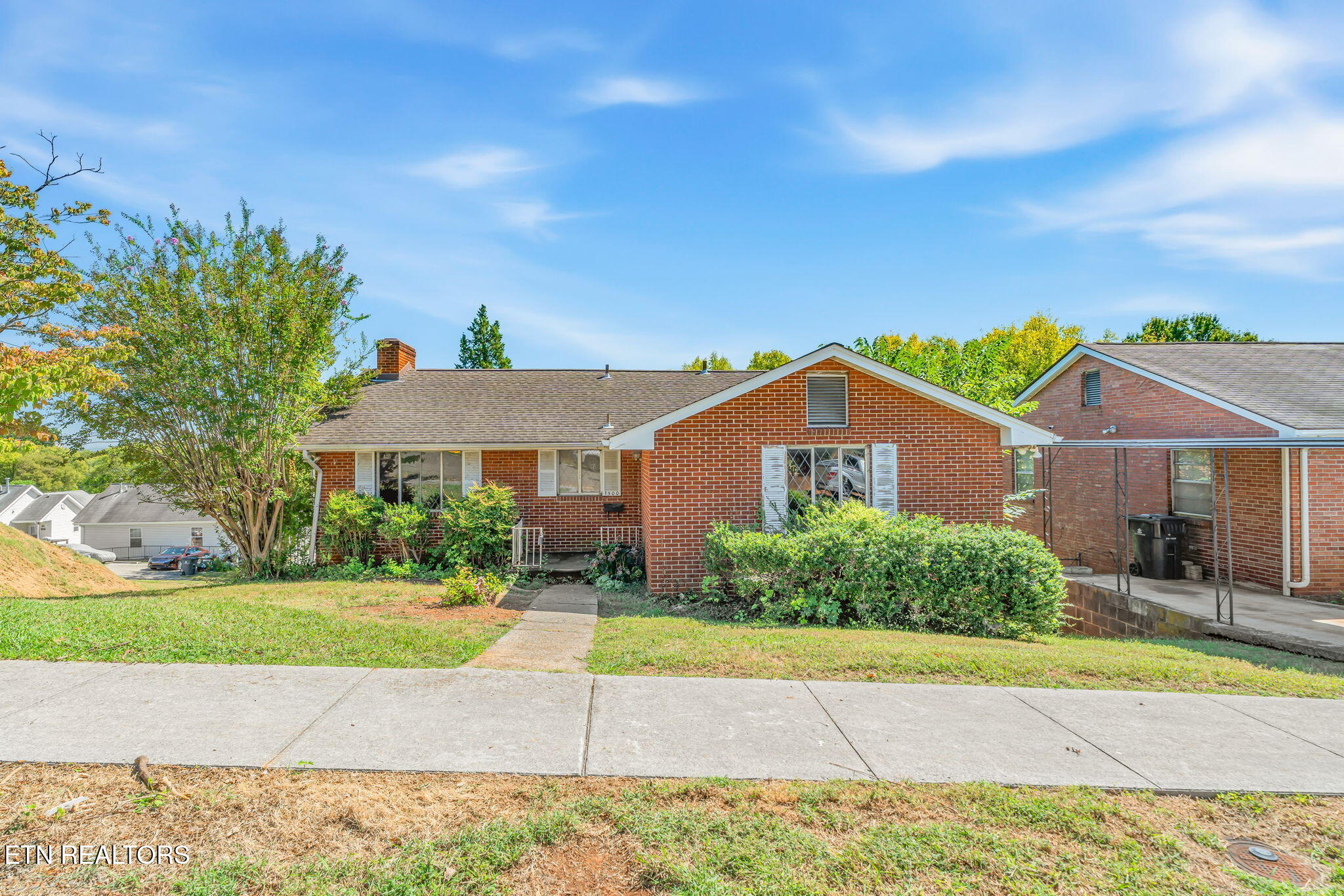 Red brick home with a gabled roof, front shrubs, and a walkway from the sidewalk.