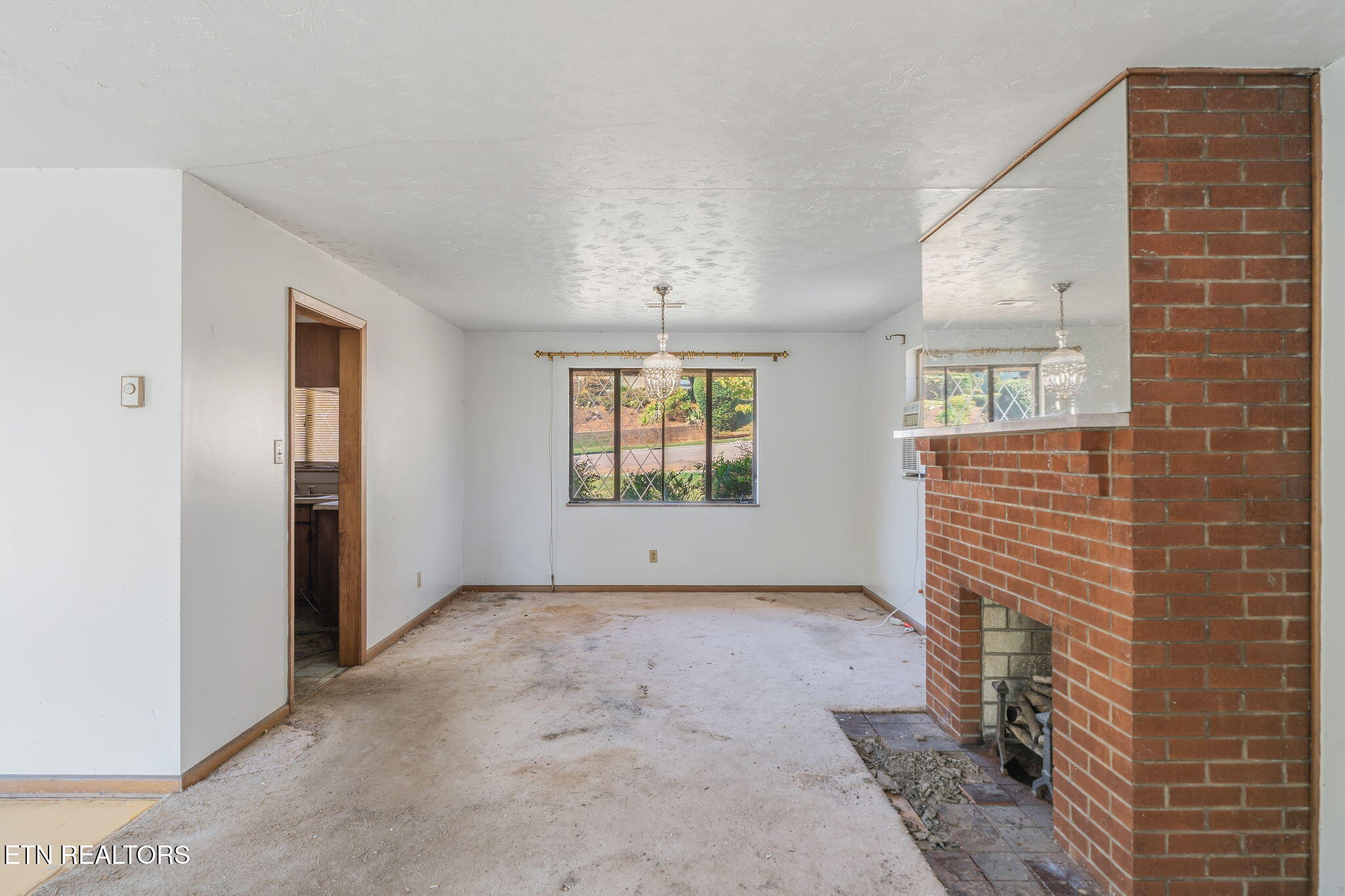 Empty living room with a large brick fireplace, mirrored mantel, and window overlooking greenery.