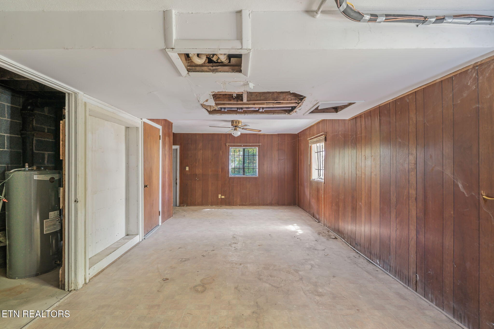 Vacant wood-paneled garage with a water heater and damaged ceiling exposing pipes and framing.
