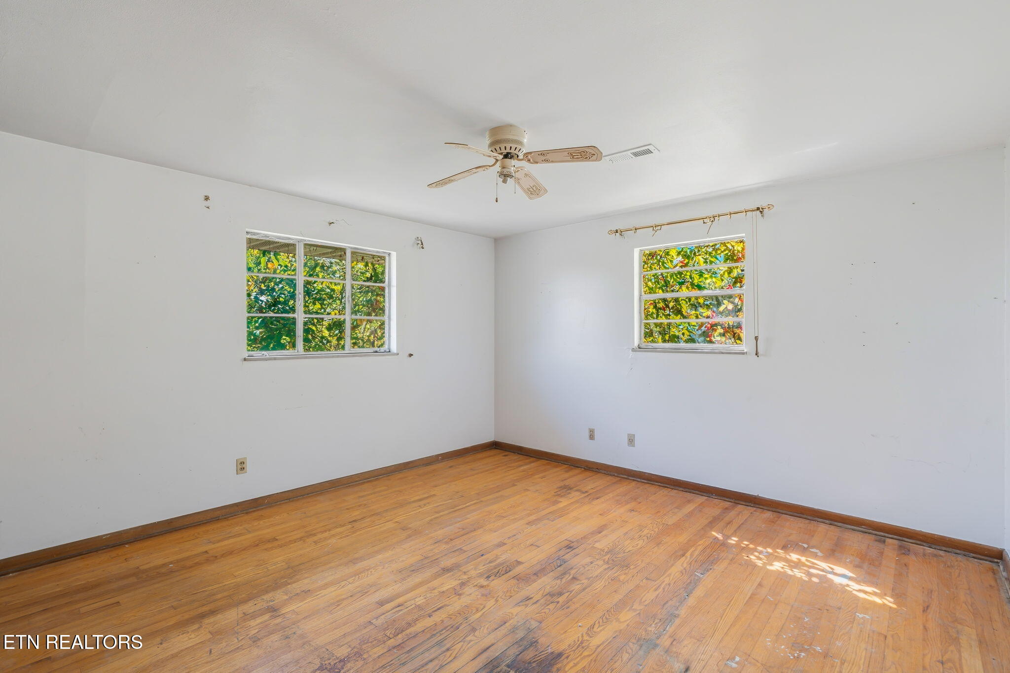 Vacant room with hardwood floors, two windows, and a ceiling fan.