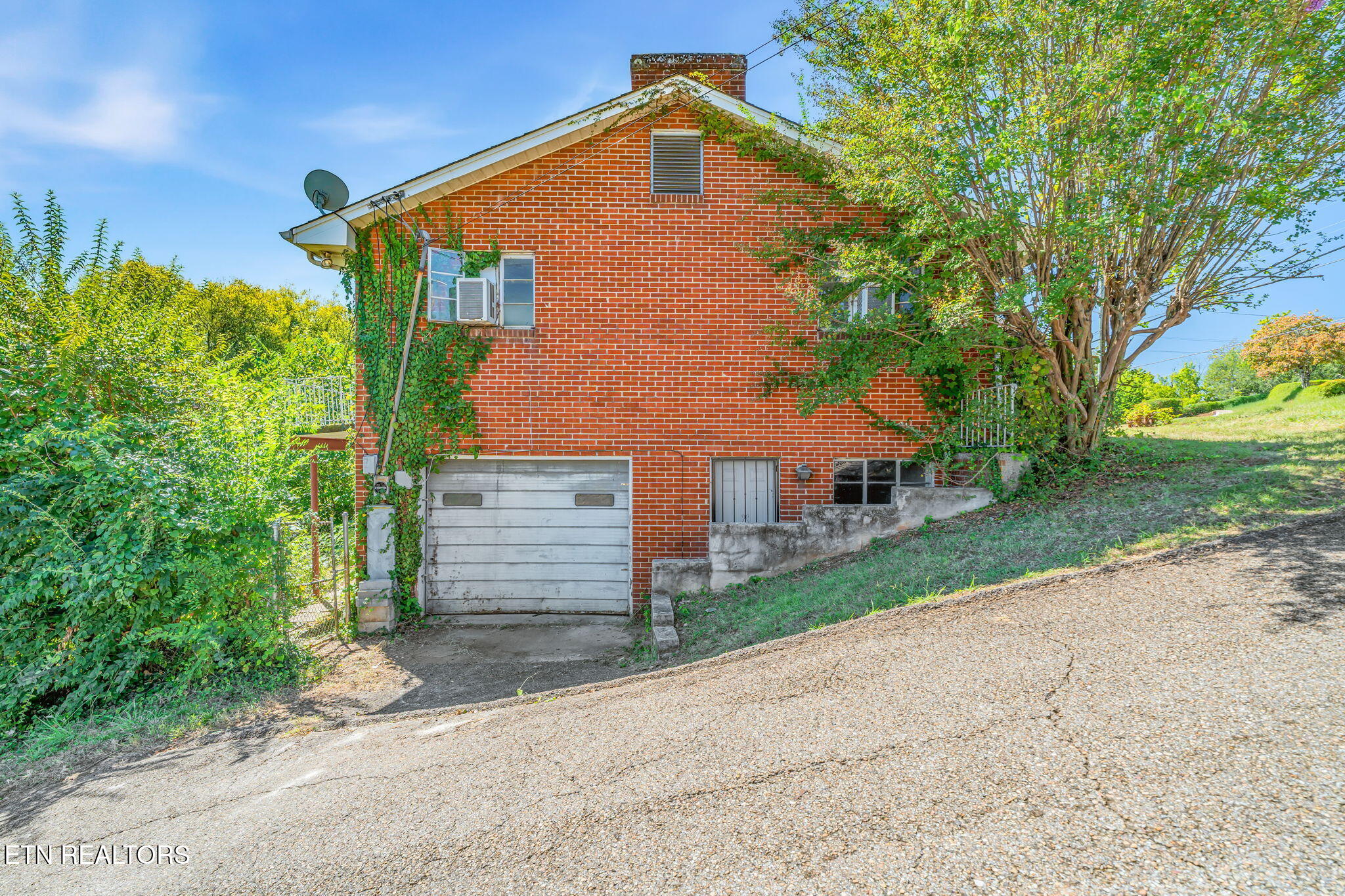 Red brick house with a garage and steep paved driveway, partly screened by trees.