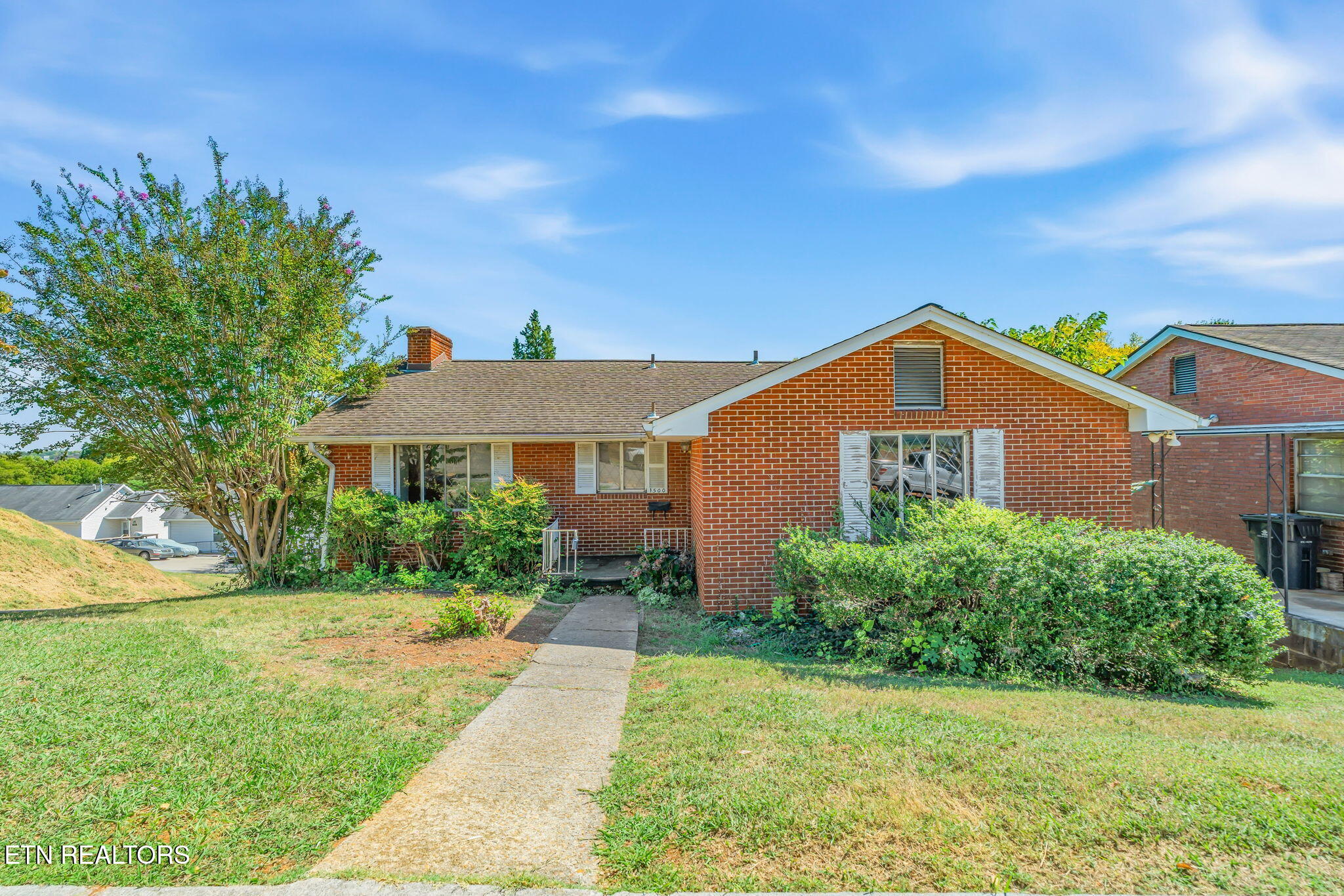 Red brick home with white-trimmed front gable, mature tree, shrubs, and a concrete walkway.