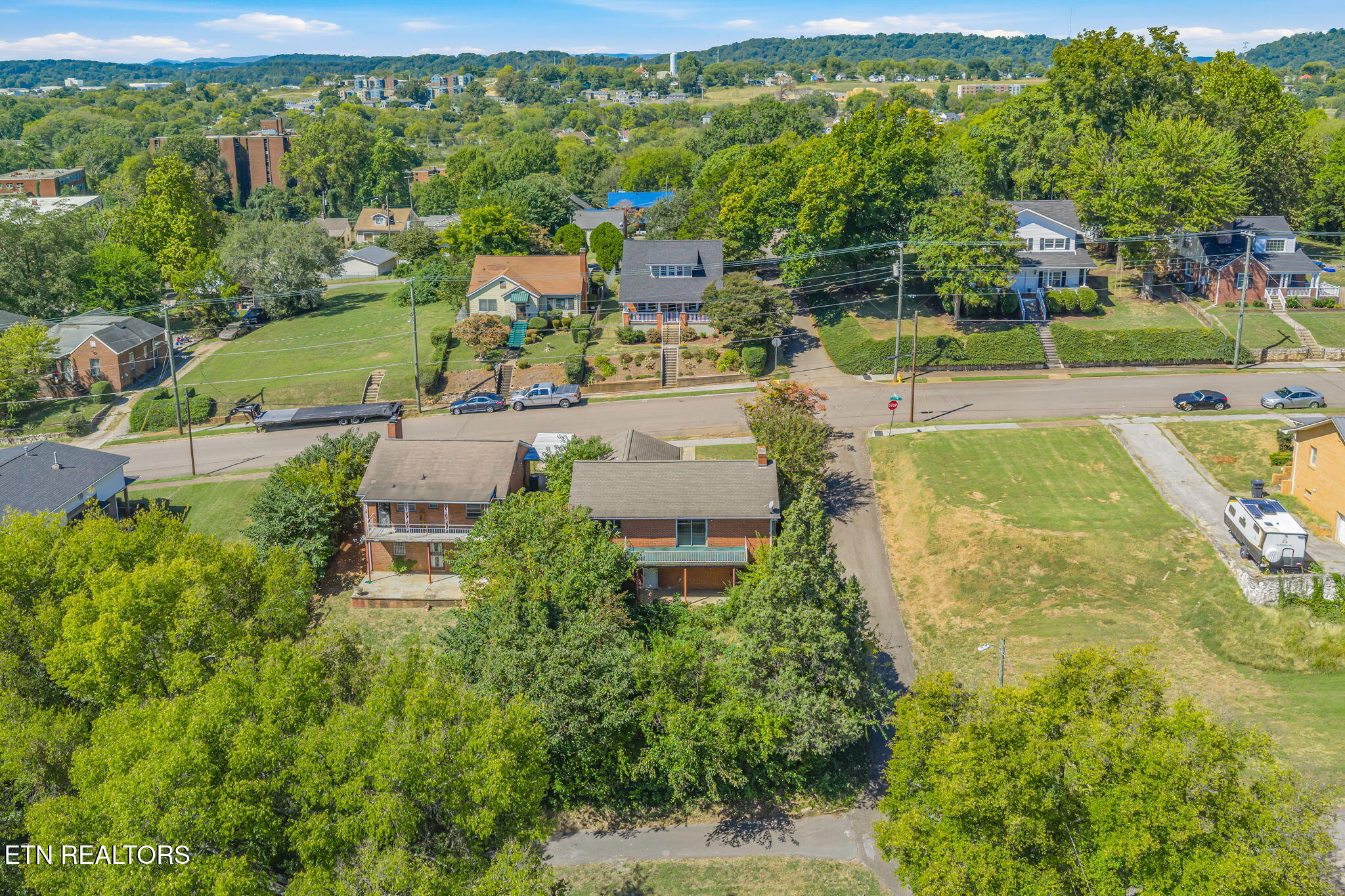 Aerial view of a multi-story brick home among leafy neighborhood homes, with wooded hills beyond.