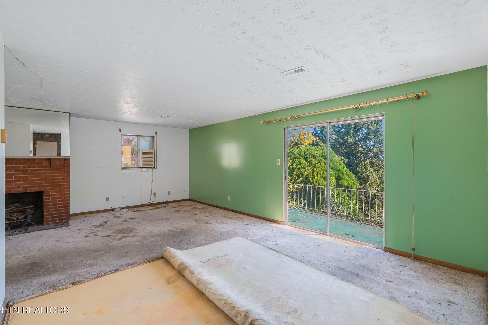 Empty living room with brick fireplace, green wall, and sliding doors to a tree-lined balcony.