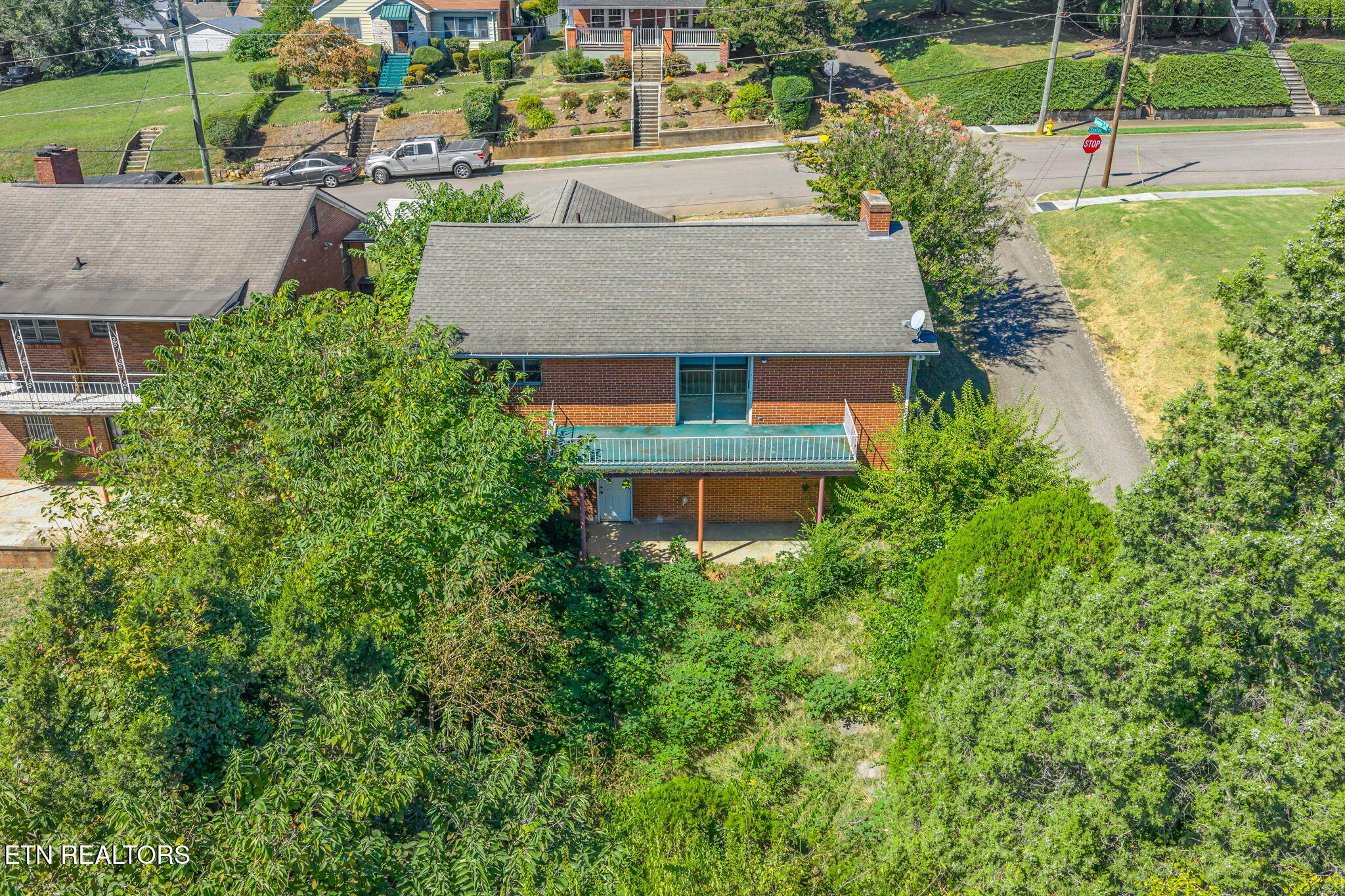 Aerial view of a brick house with a rear balcony, covered patio, and densely wooded yard.