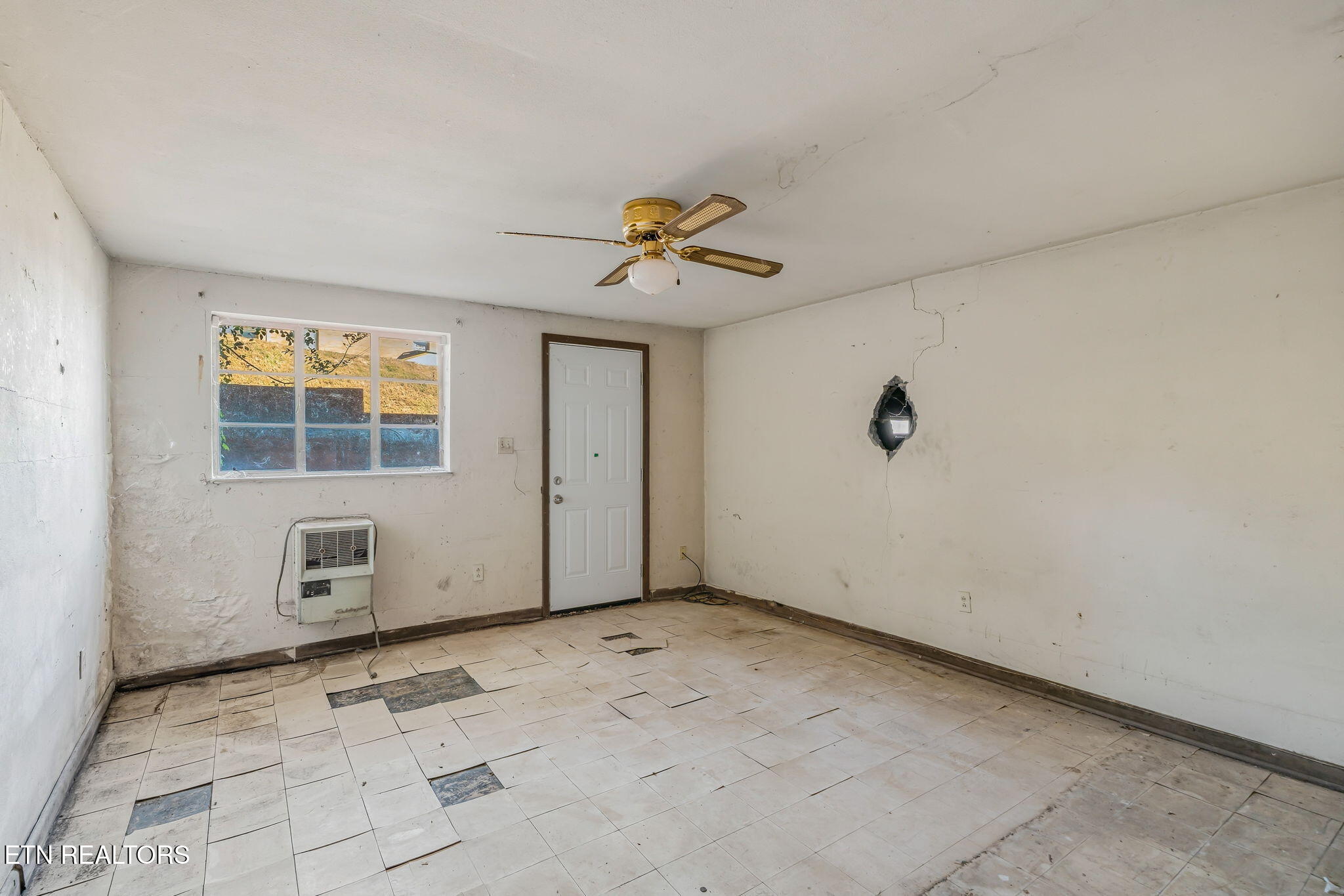 Vacant room with damaged tile flooring, cracked walls, a window, wall heater, and ceiling fan.