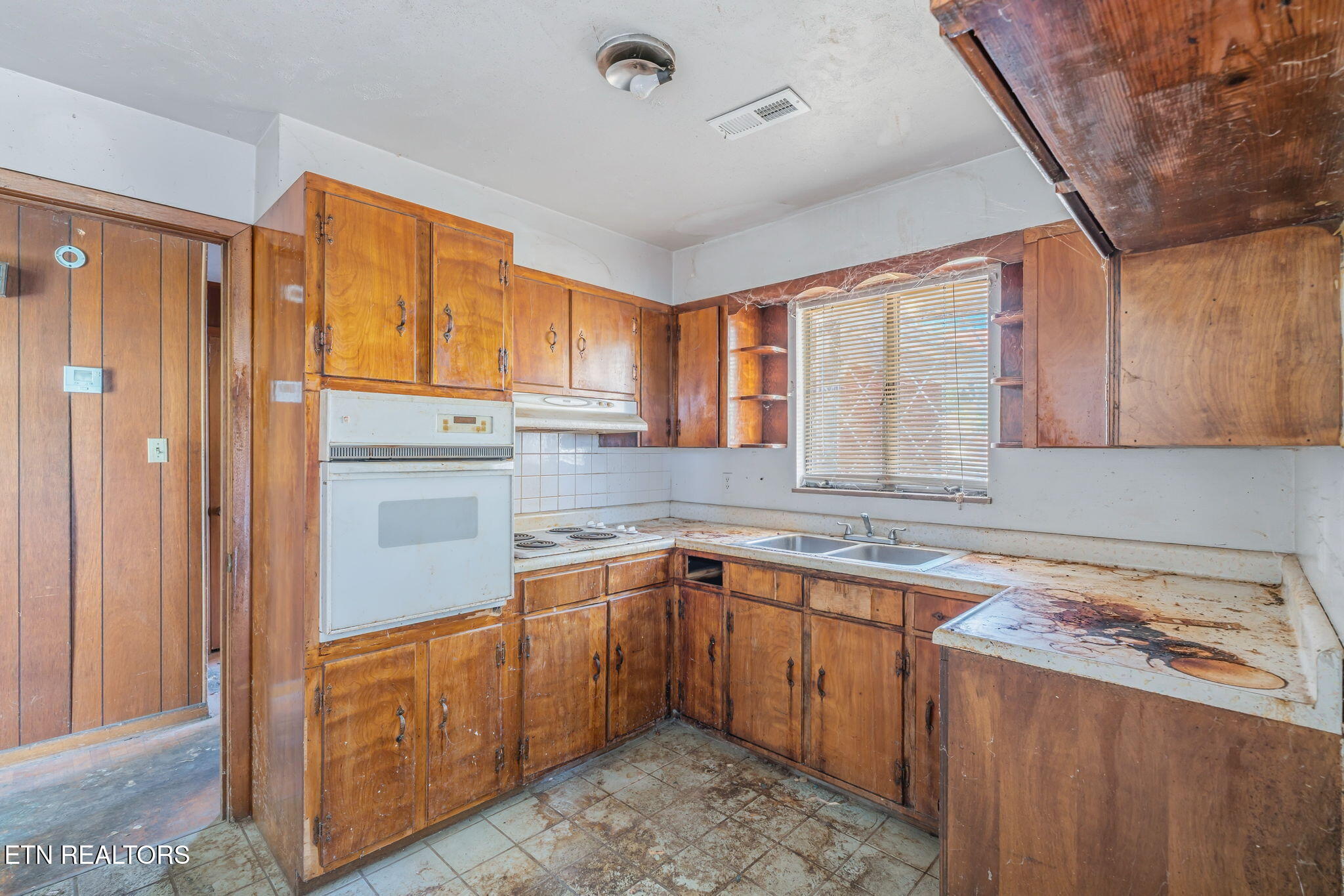 Kitchen with worn wood cabinets, an older wall oven, and a double sink beneath a window.