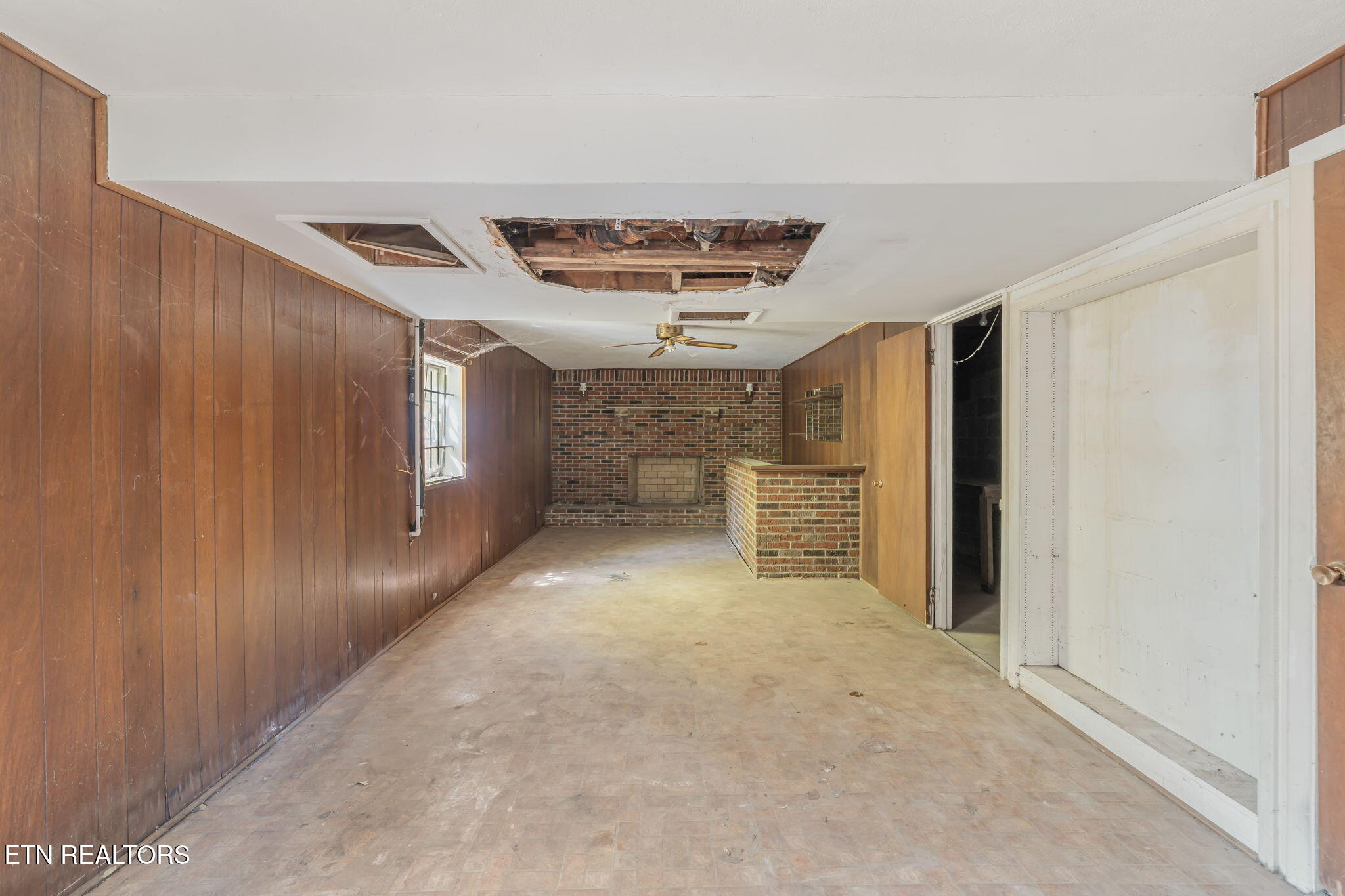 Vacant room with wood-paneled walls, brick fireplace, brick counter, and damaged open ceiling.