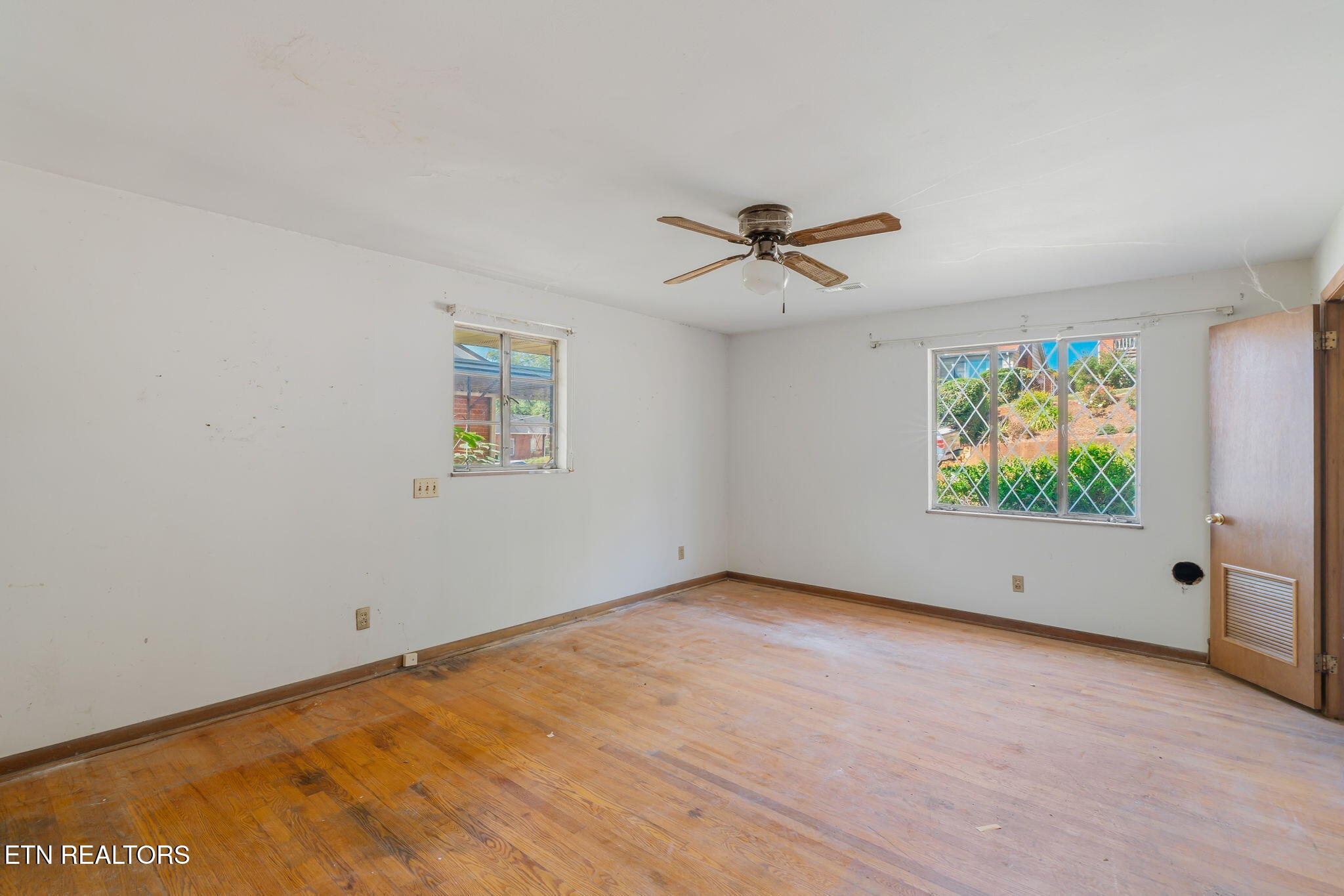 Empty room with worn hardwood floors, diamond-grille windows, and a ceiling fan.
