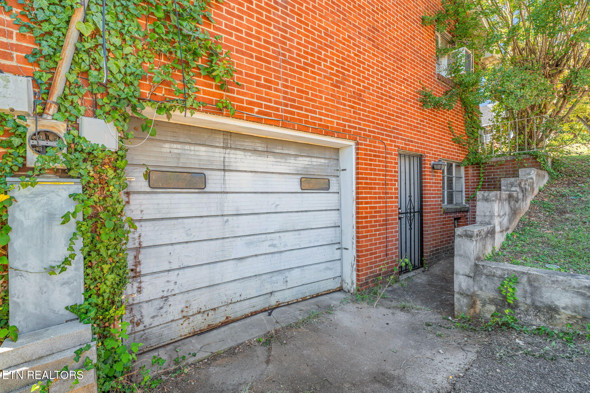 Red brick exterior with a weathered white garage door, ivy-covered wall, and barred side entrance.