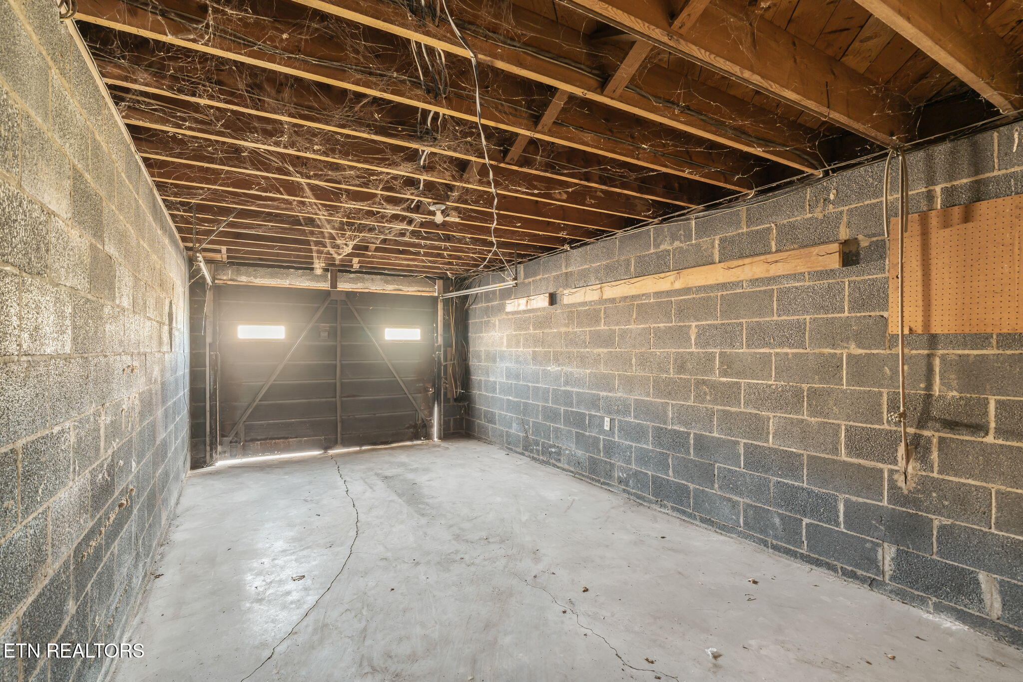 Empty garage with exposed ceiling joists, concrete block walls, and a cracked concrete floor.
