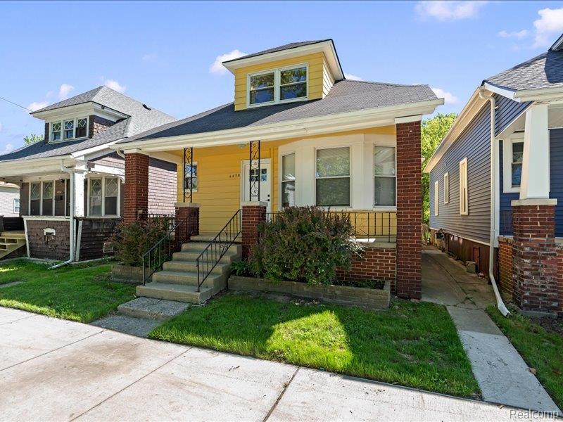 Yellow bungalow with a raised dormer, brick porch supports, and front steps.
