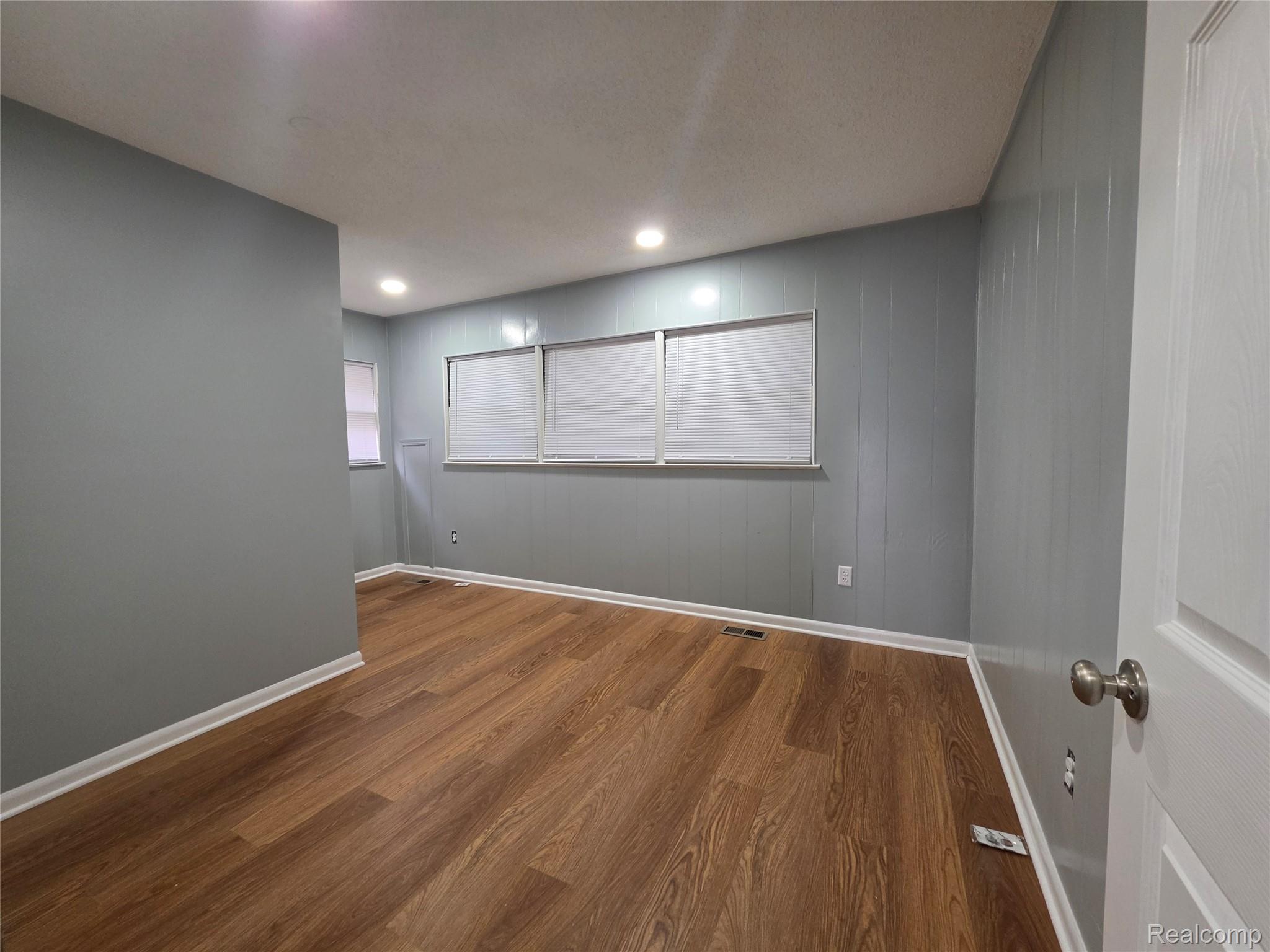 Empty room with gray paneled walls, wood-look flooring, and windows with closed blinds.