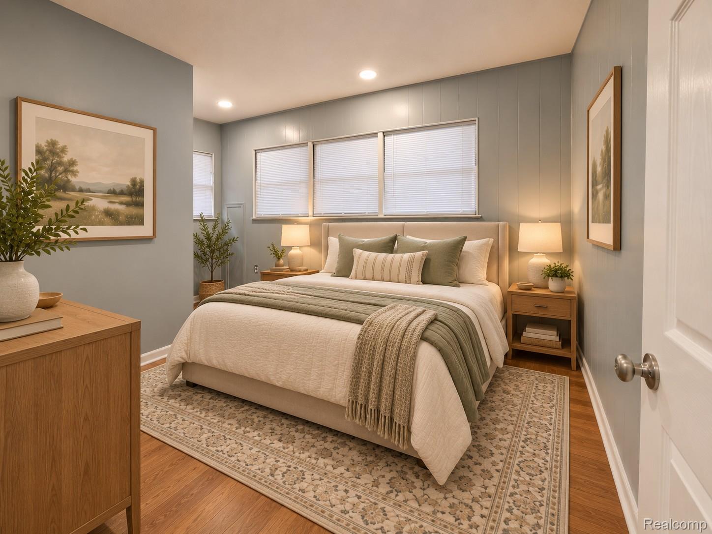 Staged bedroom with a white-and-sage bed, patterned rug, and three windows with blinds.