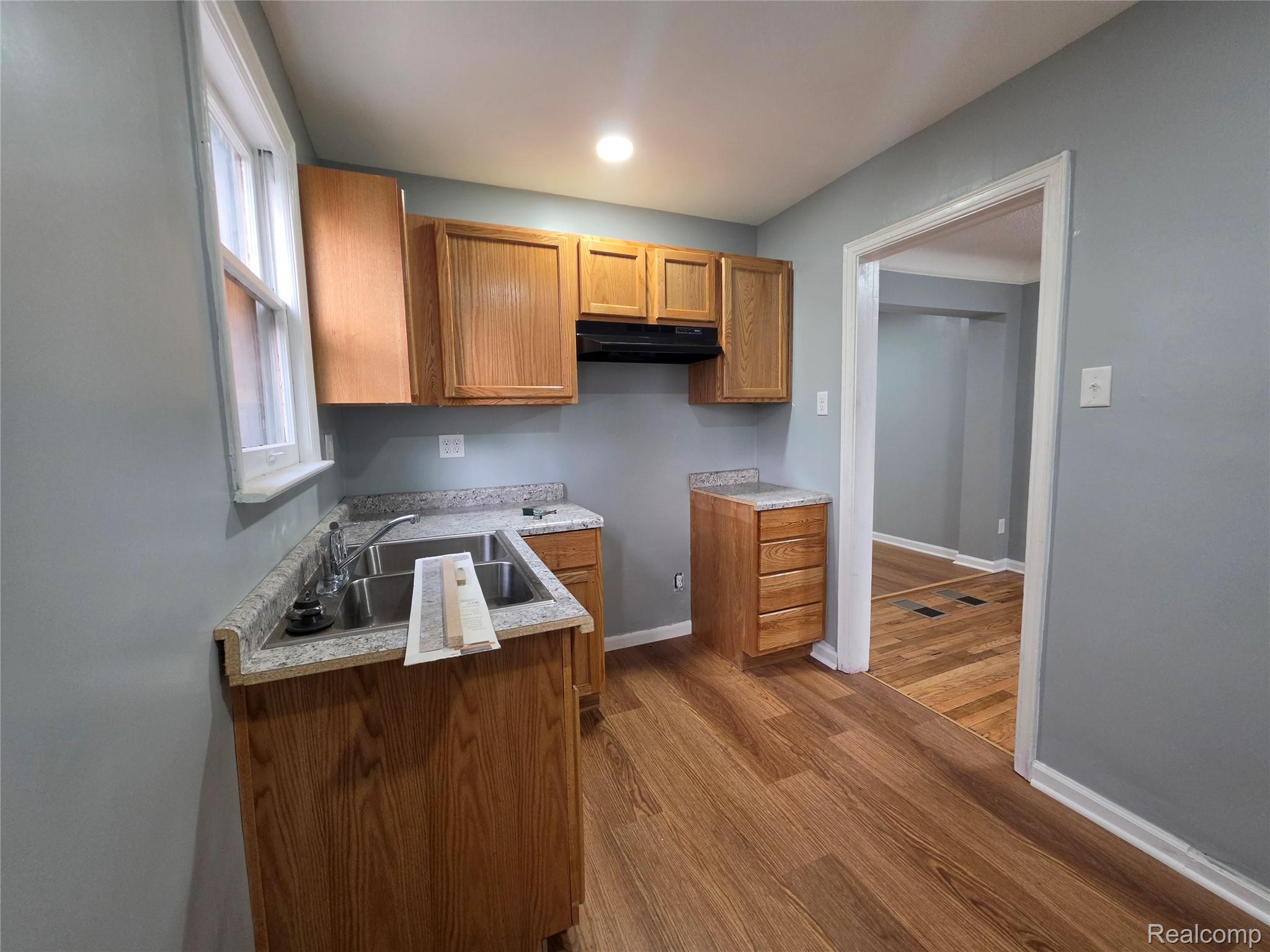 Kitchen with oak cabinets, speckled countertops, a double sink, and an open doorway to a hardwood-floored room.