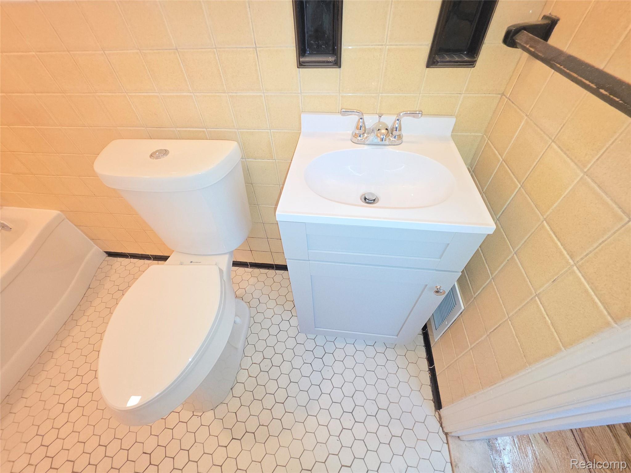 Bathroom with white toilet and vanity, tan tiled walls, and white hexagonal floor tiles.
