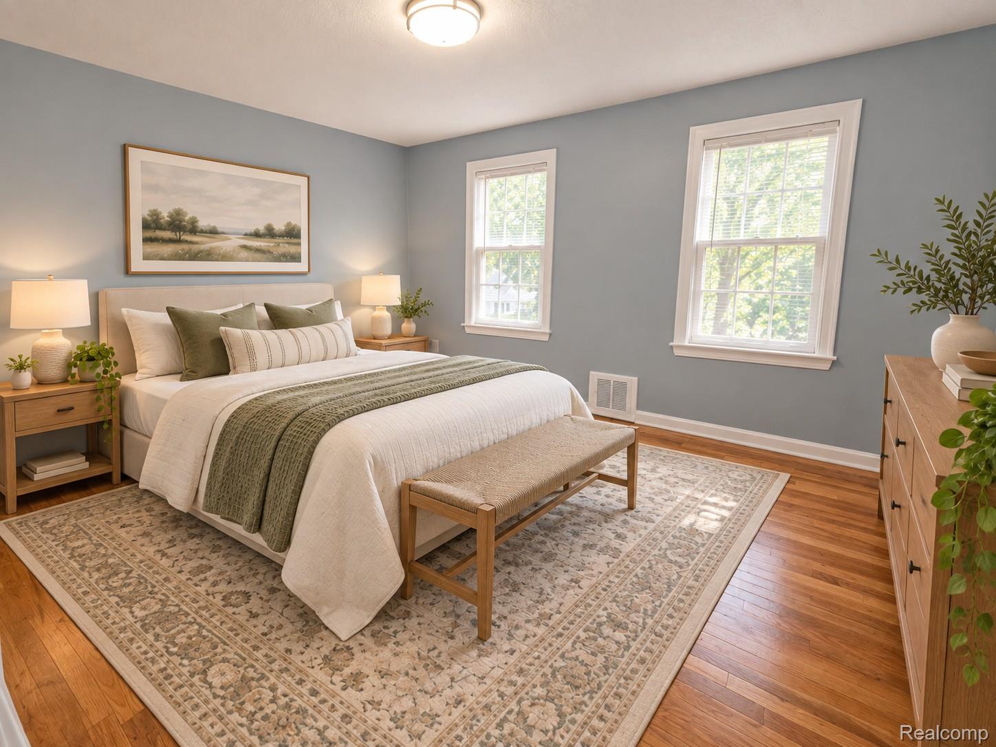 Staged bedroom with sage and cream bedding, a padded bench, and two bright windows.