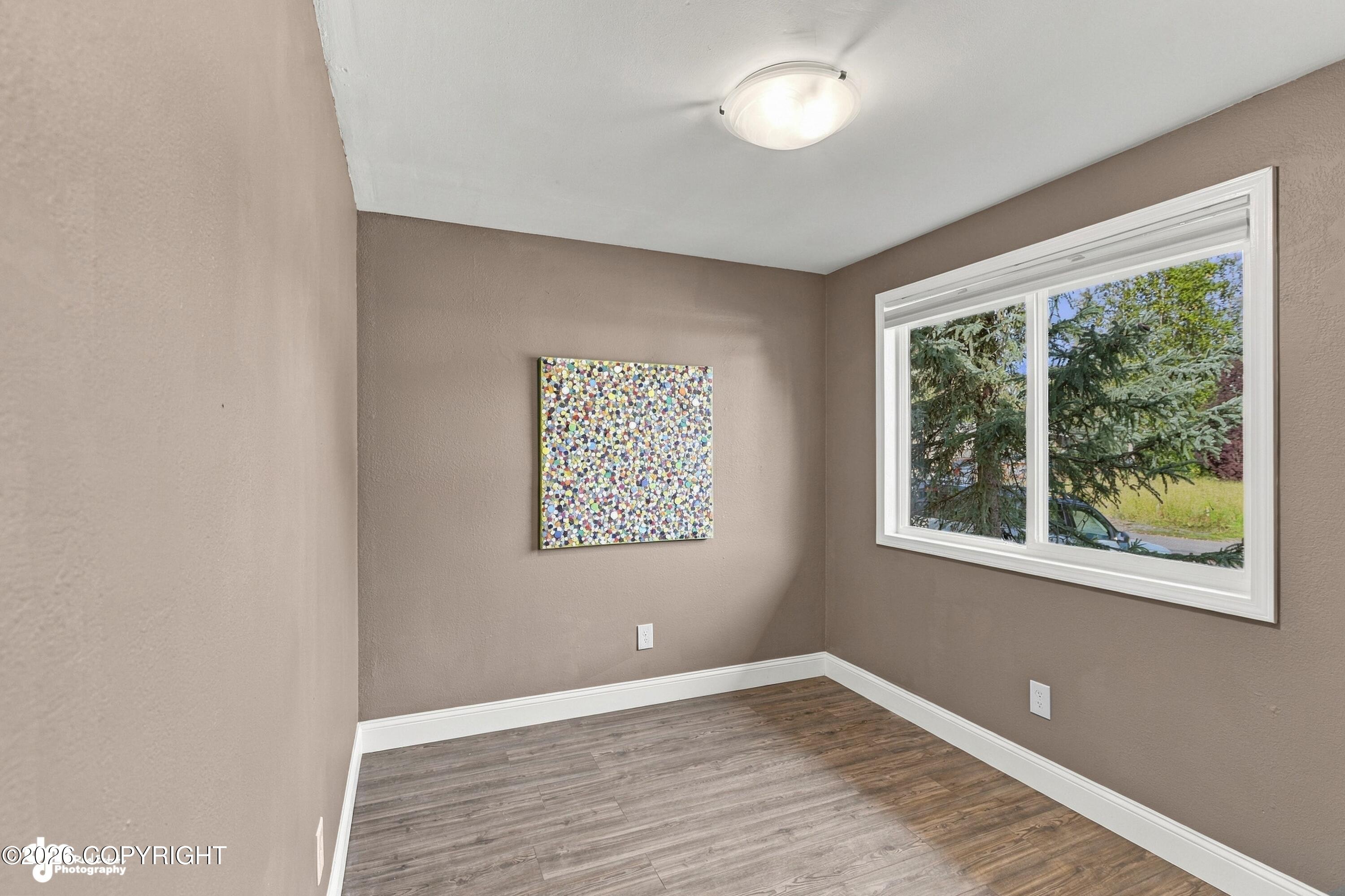 Empty room with taupe walls, wood-look flooring, and a large window overlooking trees.