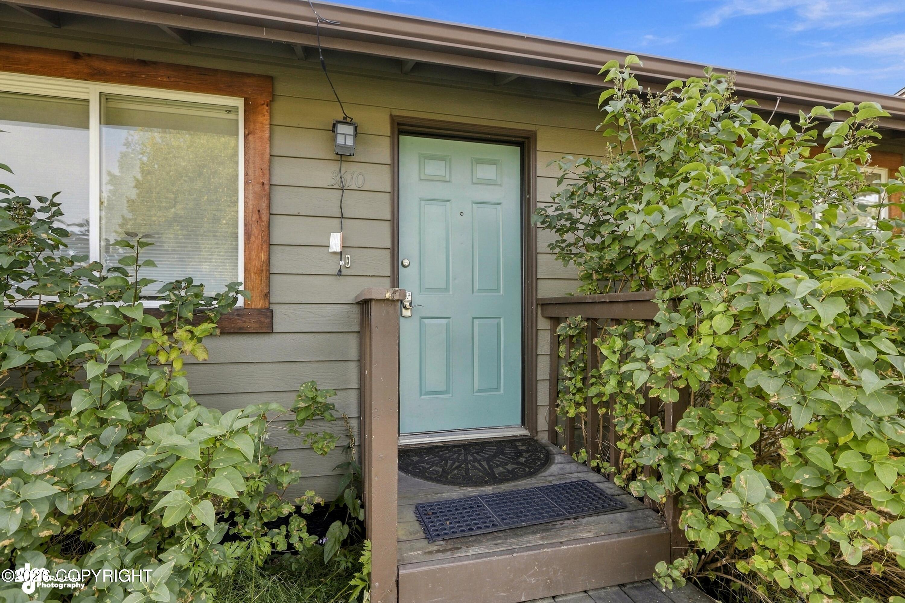 Home entrance with a teal front door, wooden porch, and leafy shrubs.