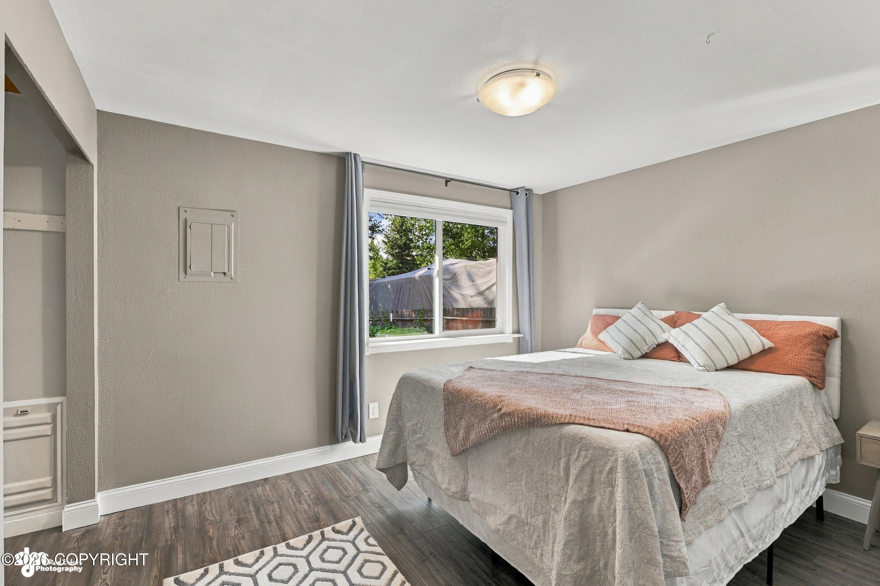 Bedroom with gray bedding, coral pillows, wood-look floors, and a window overlooking trees.