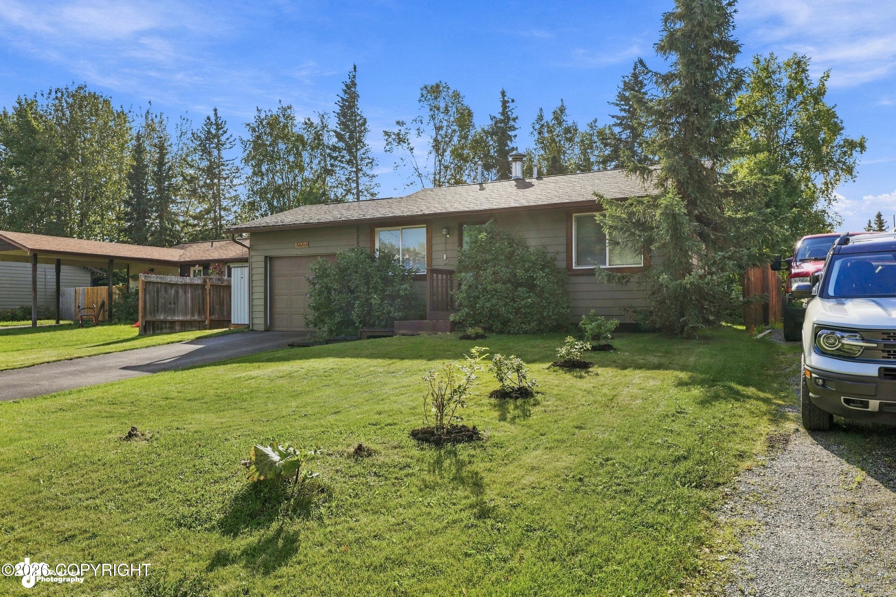 Single-story home with an attached garage, broad lawn, mature trees, and vehicles beside the driveway.