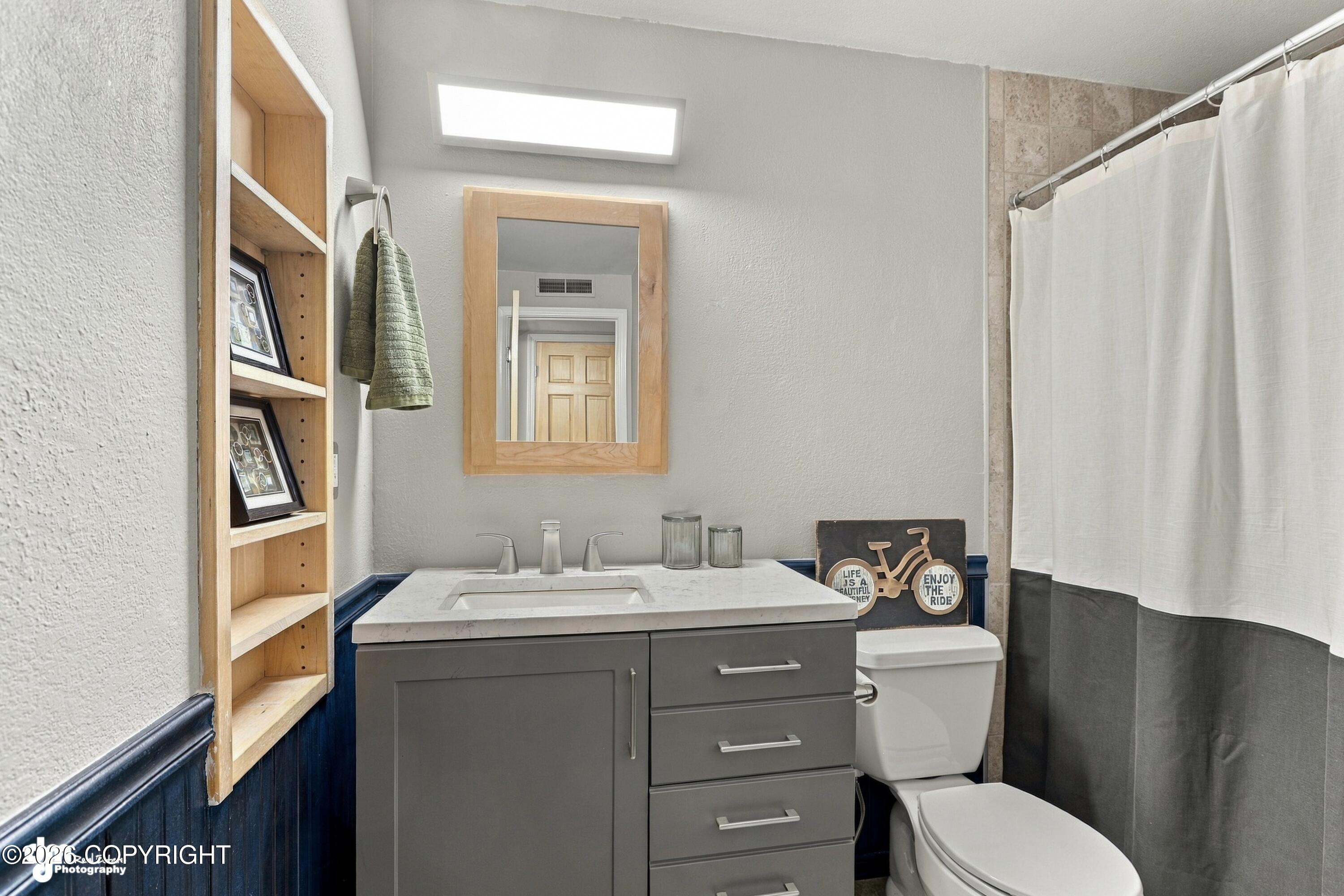 Bathroom with gray vanity, wood-framed mirror, built-in shelving, and a two-tone shower curtain.