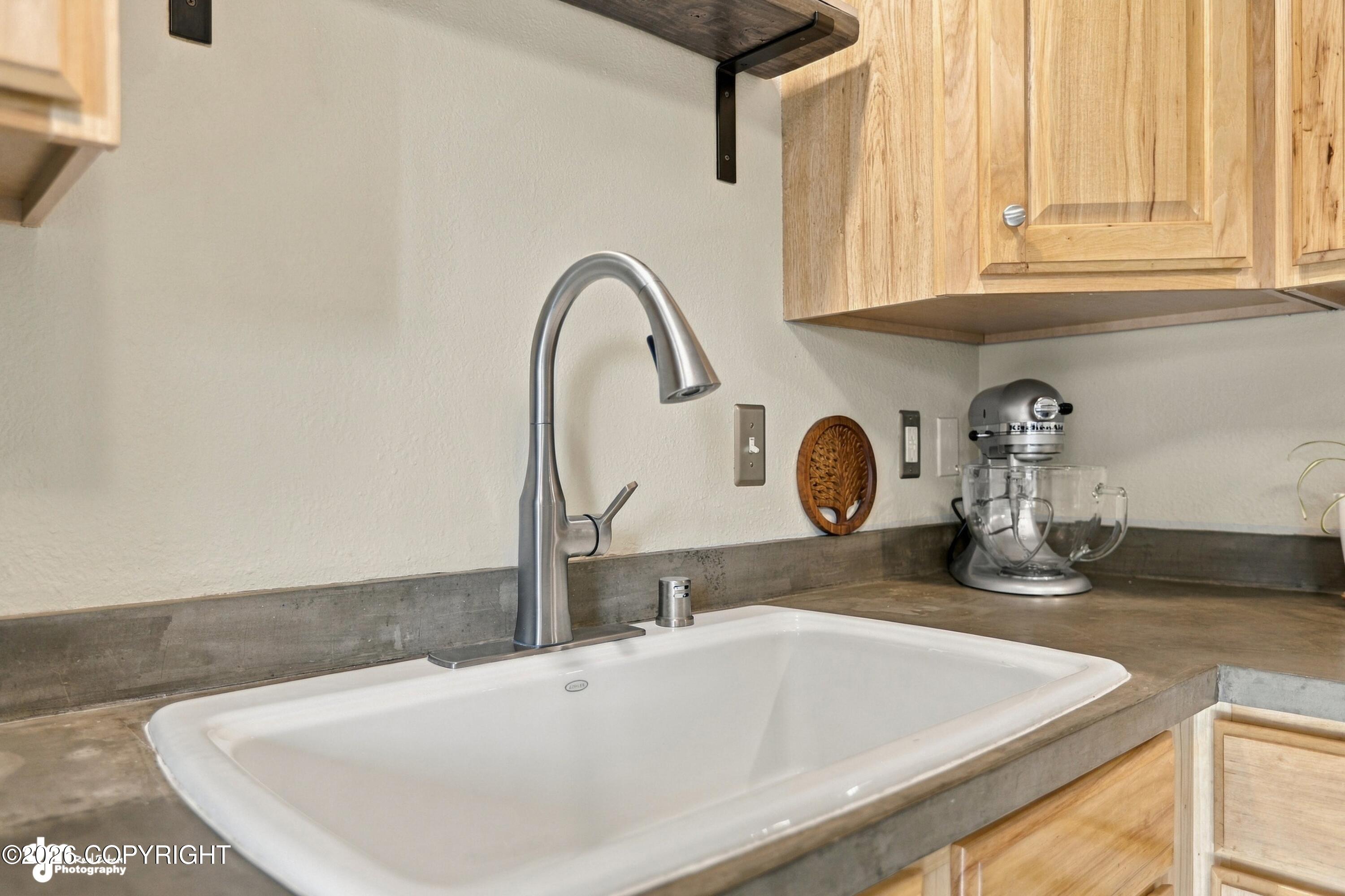 Kitchen sink with a tall stainless-steel faucet, concrete counters, light wood cabinets, and a stand mixer.