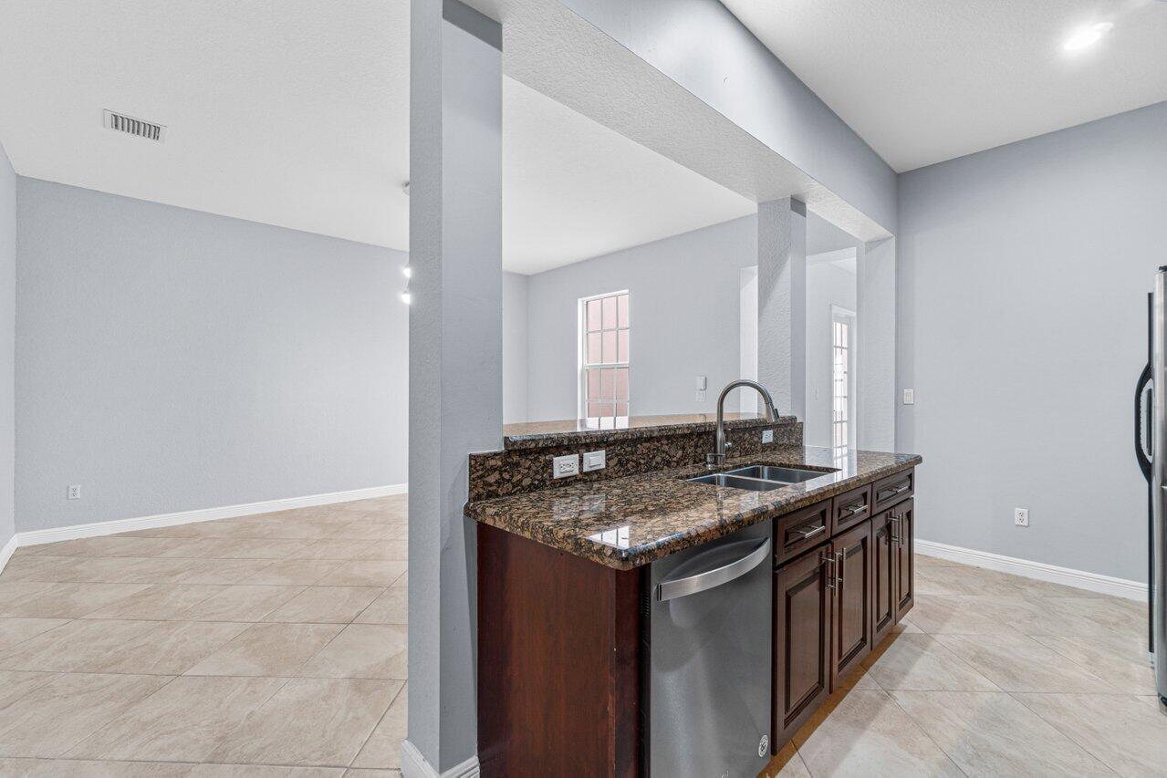 Open kitchen with dark wood cabinets, granite counters, stainless dishwasher, and a large tiled adjoining room.