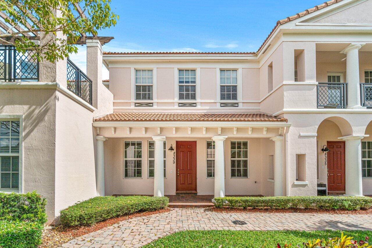 Pale stucco two-story townhouse with a red entry door, tiled roof, and landscaped front walk.