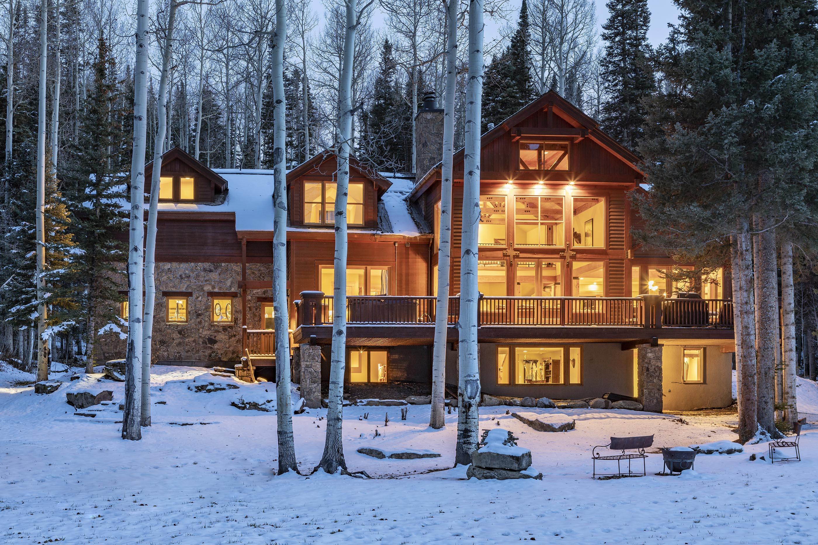 Mountain home with a broad deck, glowing windows, and snow-covered grounds among aspens.