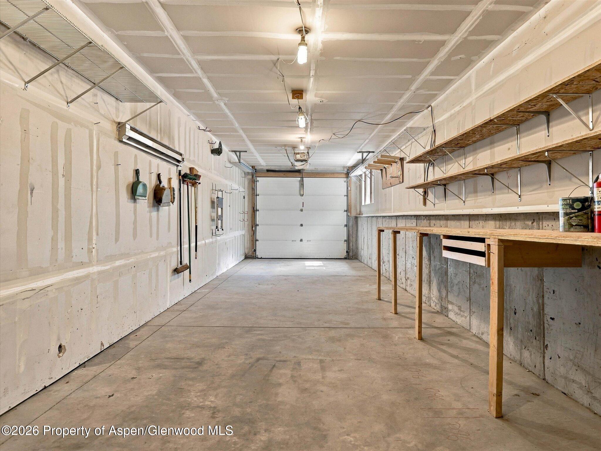 Garage with a concrete floor, overhead door, wall shelves, and a long wooden workbench.
