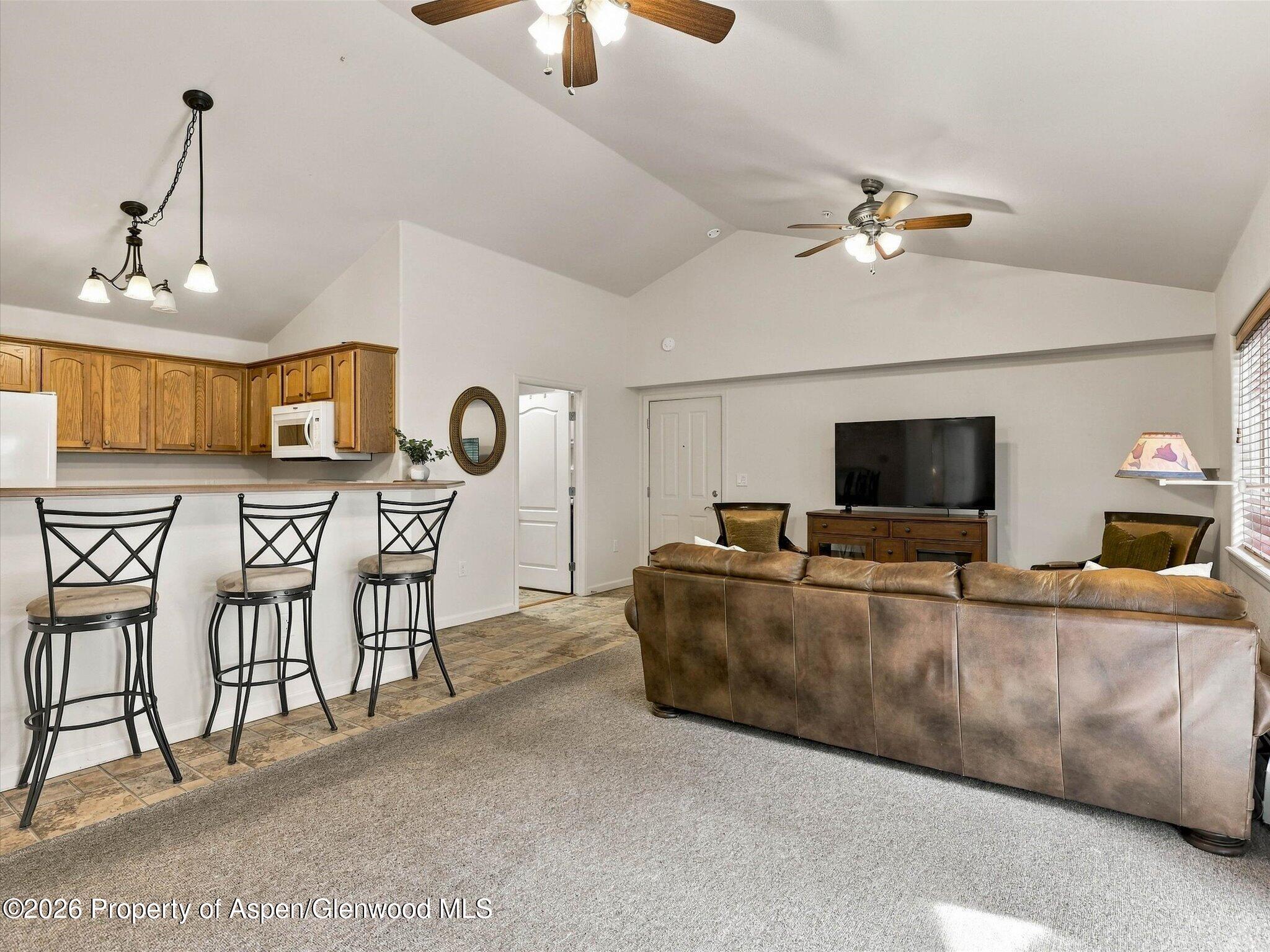 Furnished living room with a brown leather sectional, vaulted ceiling, and open kitchen breakfast bar.
