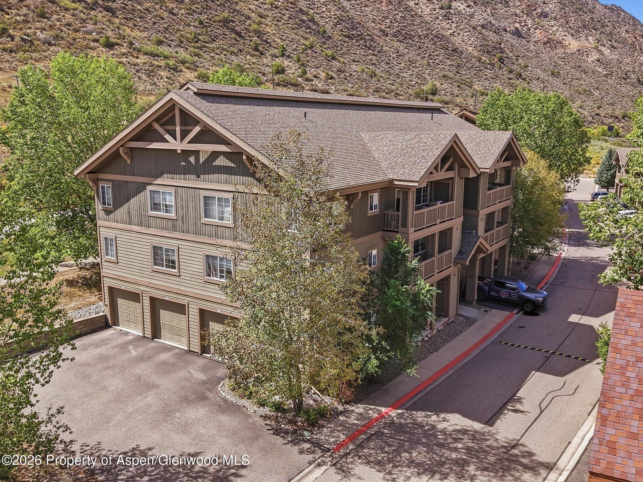 Multi-story residential building with timber-style gables, balconies, and garages beside a tree-lined drive.