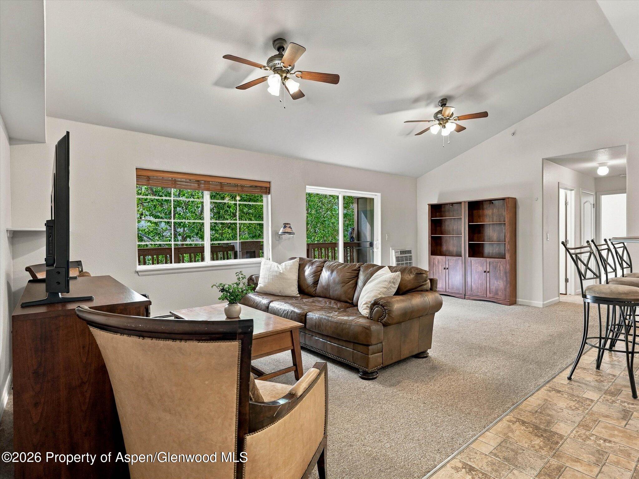 Furnished living room with a leather sofa, vaulted ceiling, ceiling fans, and windows overlooking trees.